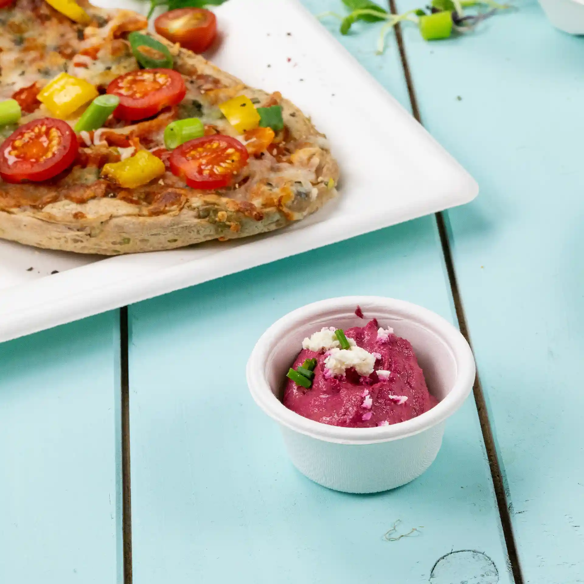 White bagasse dip bowl with pink dip and green garnish, placed beside a pizza on a white plate.