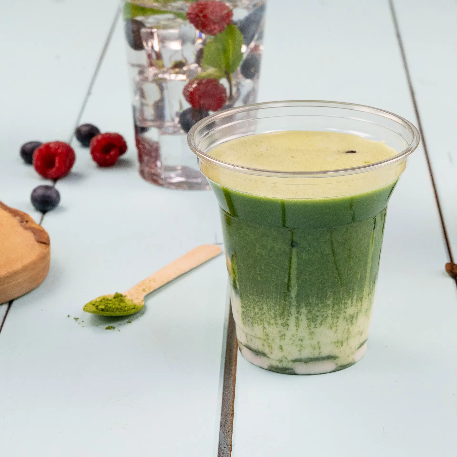 A clear 8 oz rPET disposable cup filled with a green layered beverage, placed on a light blue surface, surrounded by raspberries, blueberries, and a glass of water.