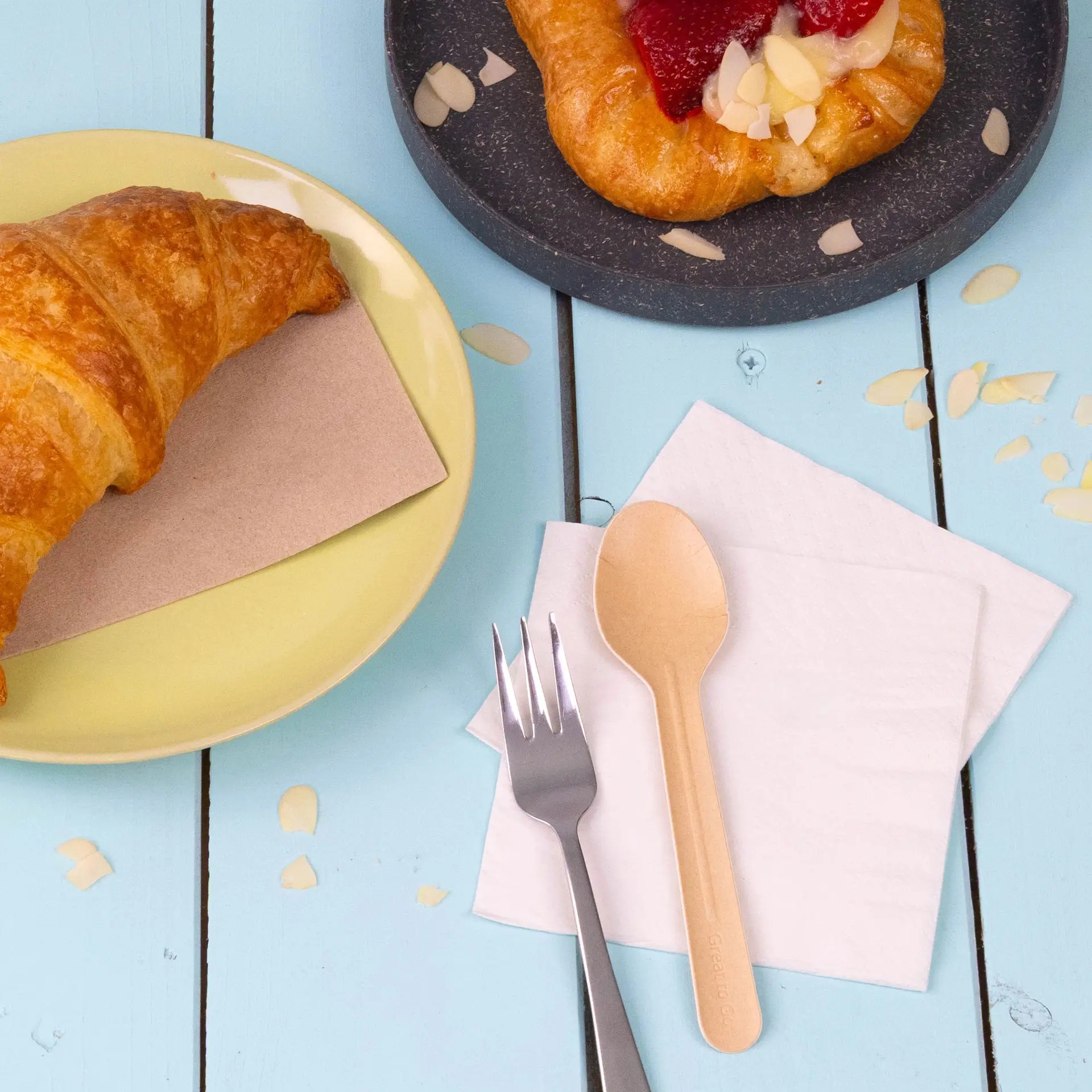 Image of a croissant on a yellow plate, a dessert on a dark plate, a wooden spoon, a silver fork, and white napkins.