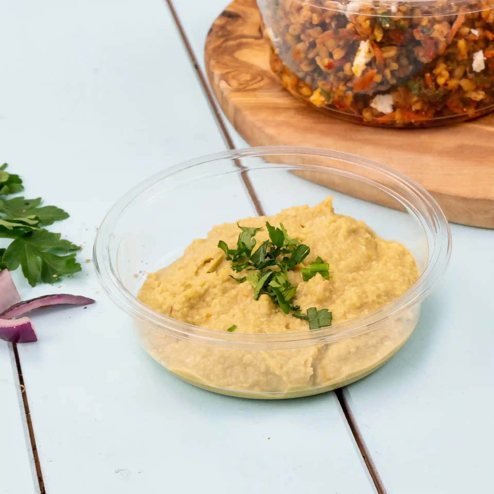 Round transparent deli container with hummus topped with chopped parsley on a light blue surface.