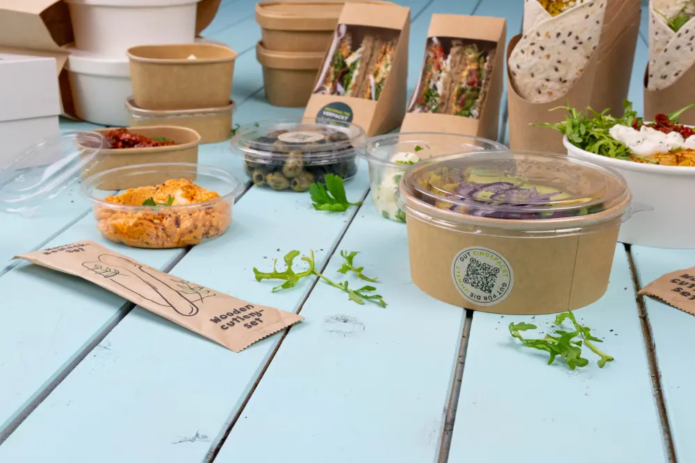 Assorted food packaging items on a blue surface, featuring salad containers, sandwich wraps, and a wooden cutlery set.