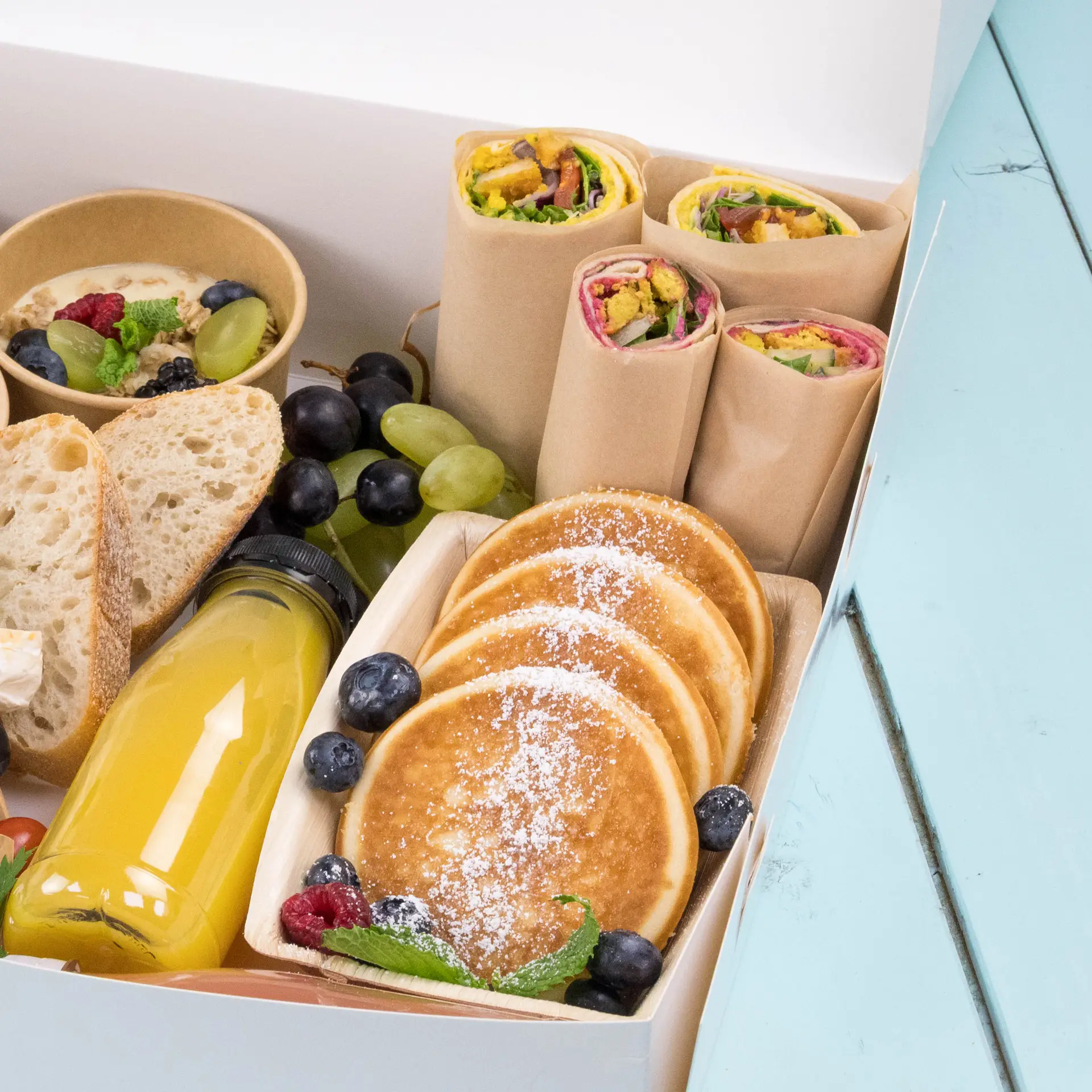 A box filled with breakfast items, including wraps, pancakes, fruit, bread, and a bottle of juice.