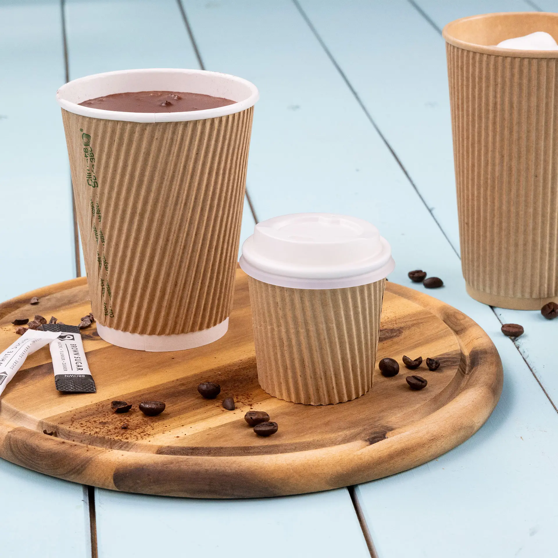 One ribbed brown coffee cup with hot chocolate, a smaller cup with a white lid, and a larger cup beside them on a wooden tray.
