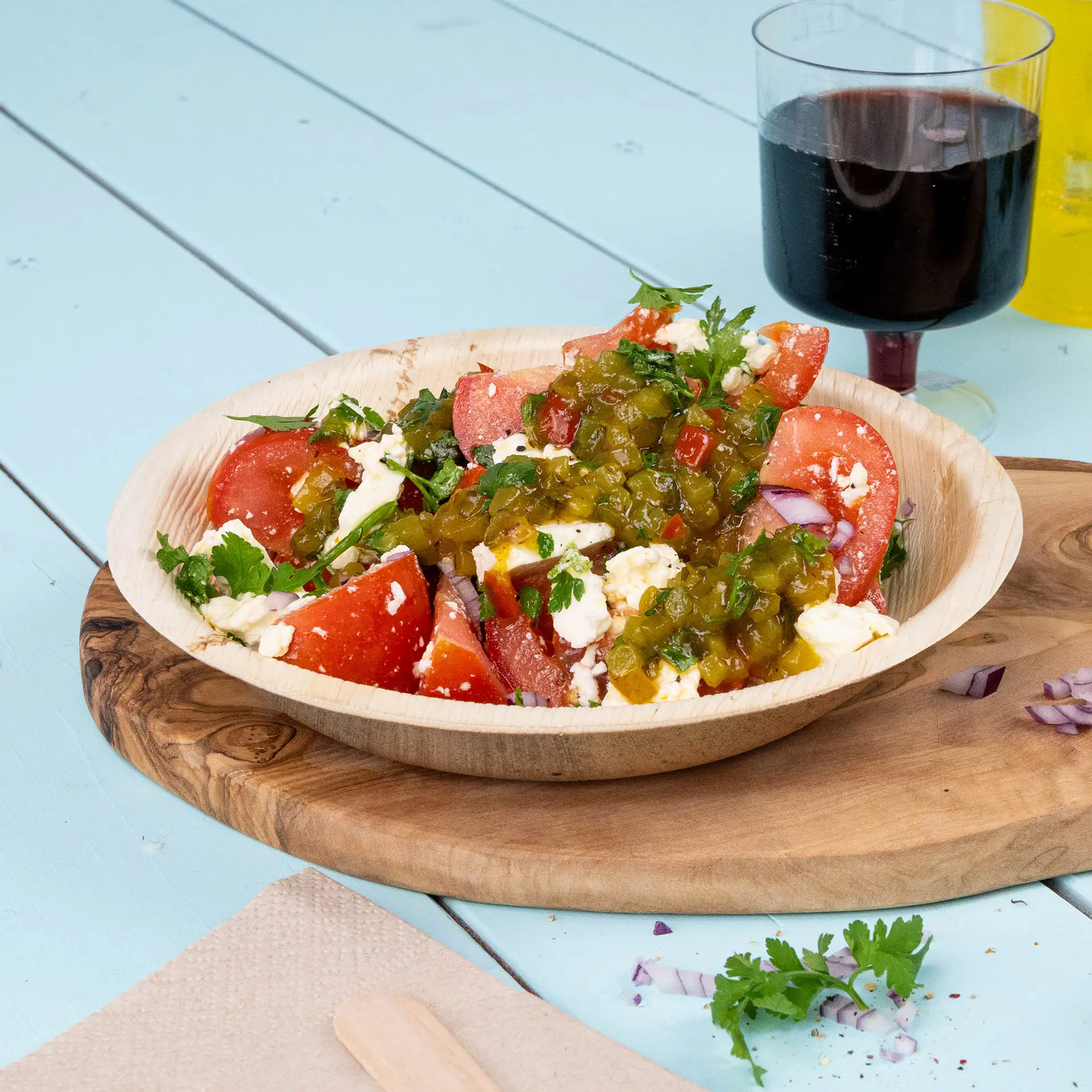 A deep round palm leaf plate filled with a salad of tomatoes, herbs, and jalapeño, set on a wooden board with a beverage.