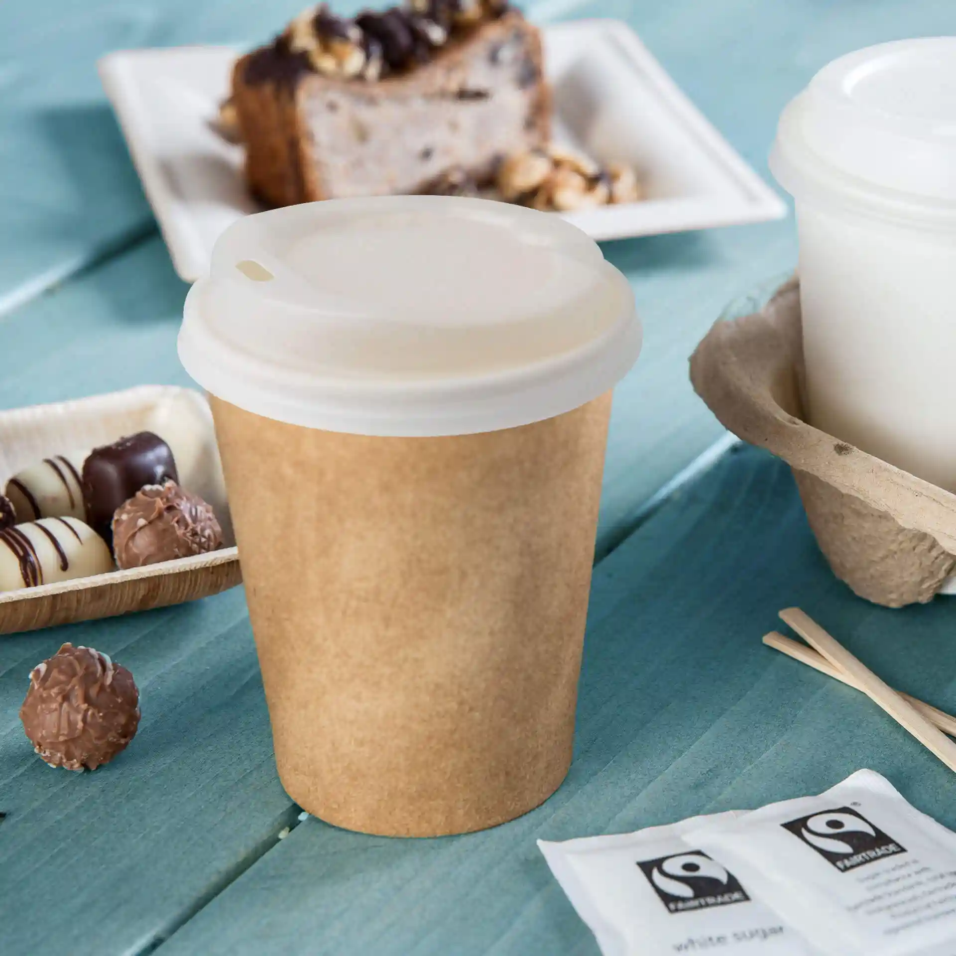 Brown paper cup with a white lid, placed on a blue surface, surrounded by chocolates and sugar packets.