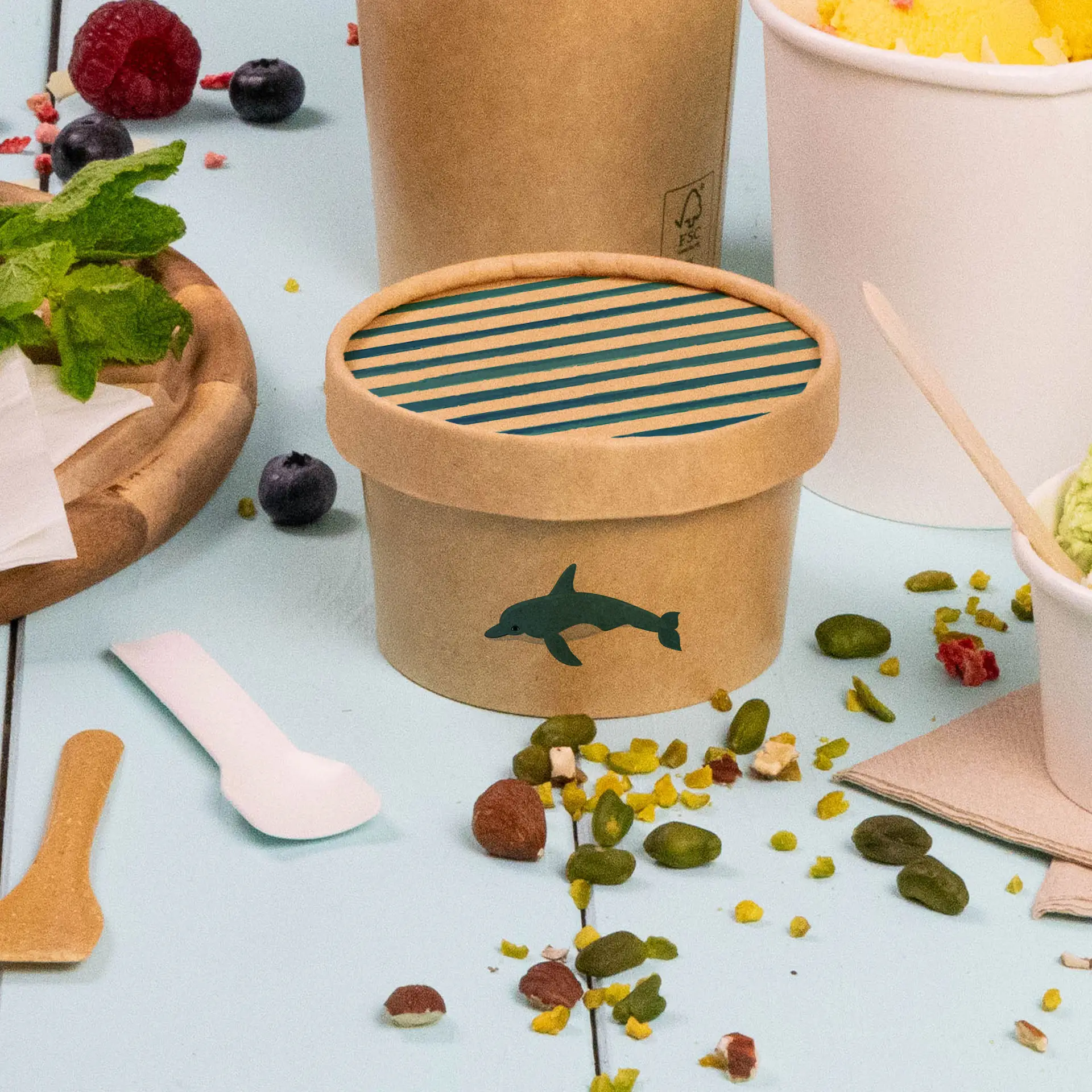 Brown cardboard lid with printed dolphin design and striped top, designed for ice cream cups, surrounded by spilled toppings.