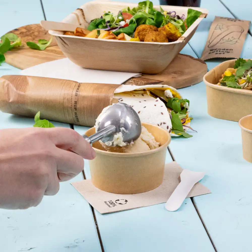 A hand using a metal ice cream scoop to serve from a brown paper cup; other food items and containers are in the background on a blue table.