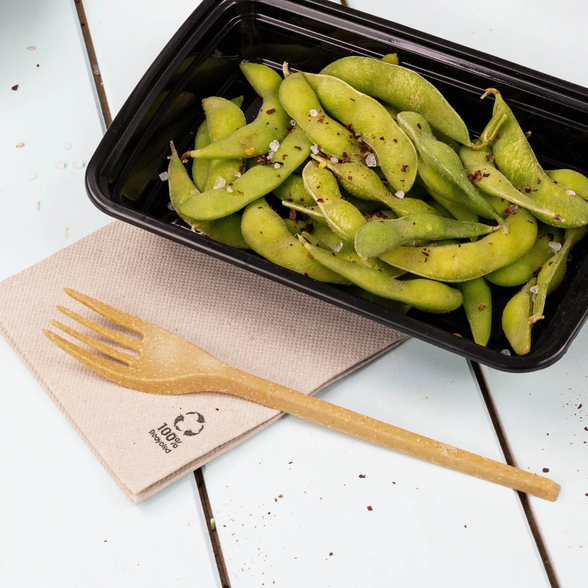 Image of fresh edamame beans in a black container, accompanied by a biodegradable fork on a recycled paper napkin.