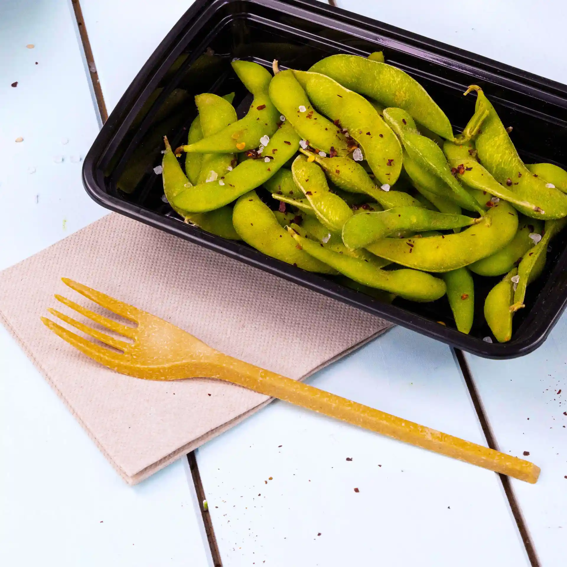 Image of a black container filled with green edamame, a yellow fork on a beige napkin beside it.