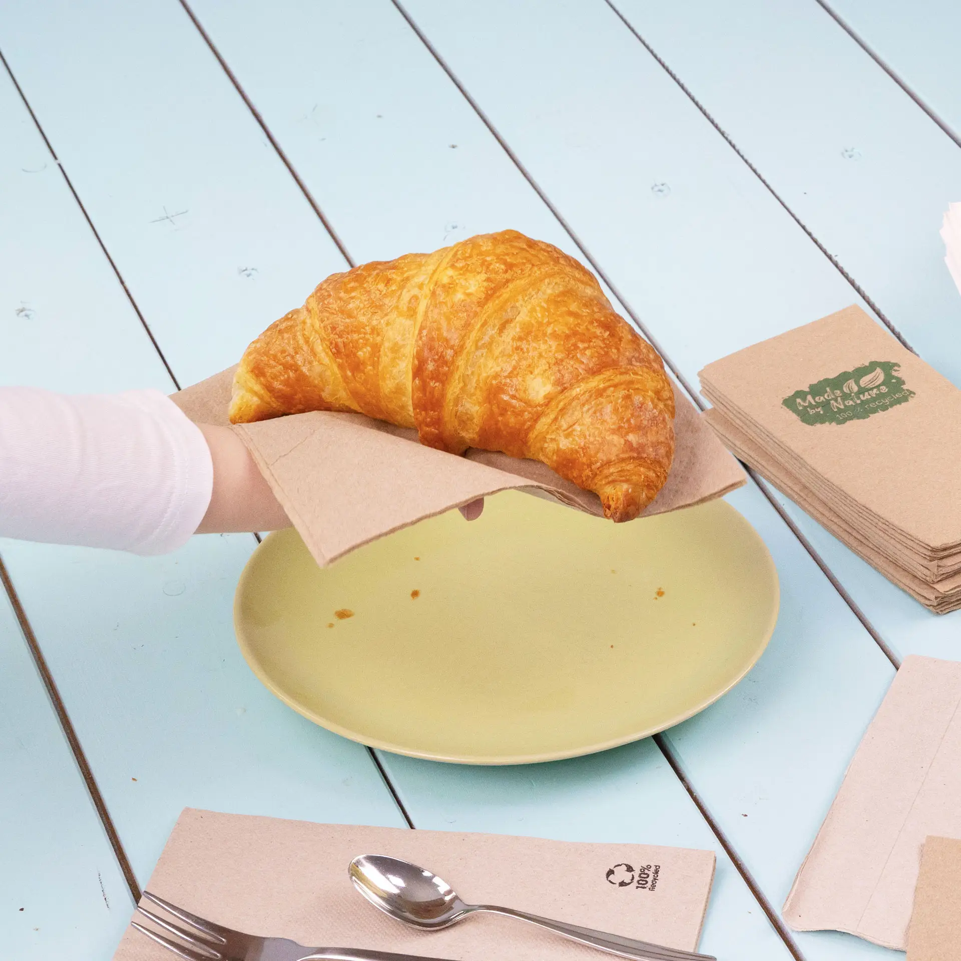 A hand holding a croissant on a brown paper napkin above a yellow plate, with additional napkins in the background.