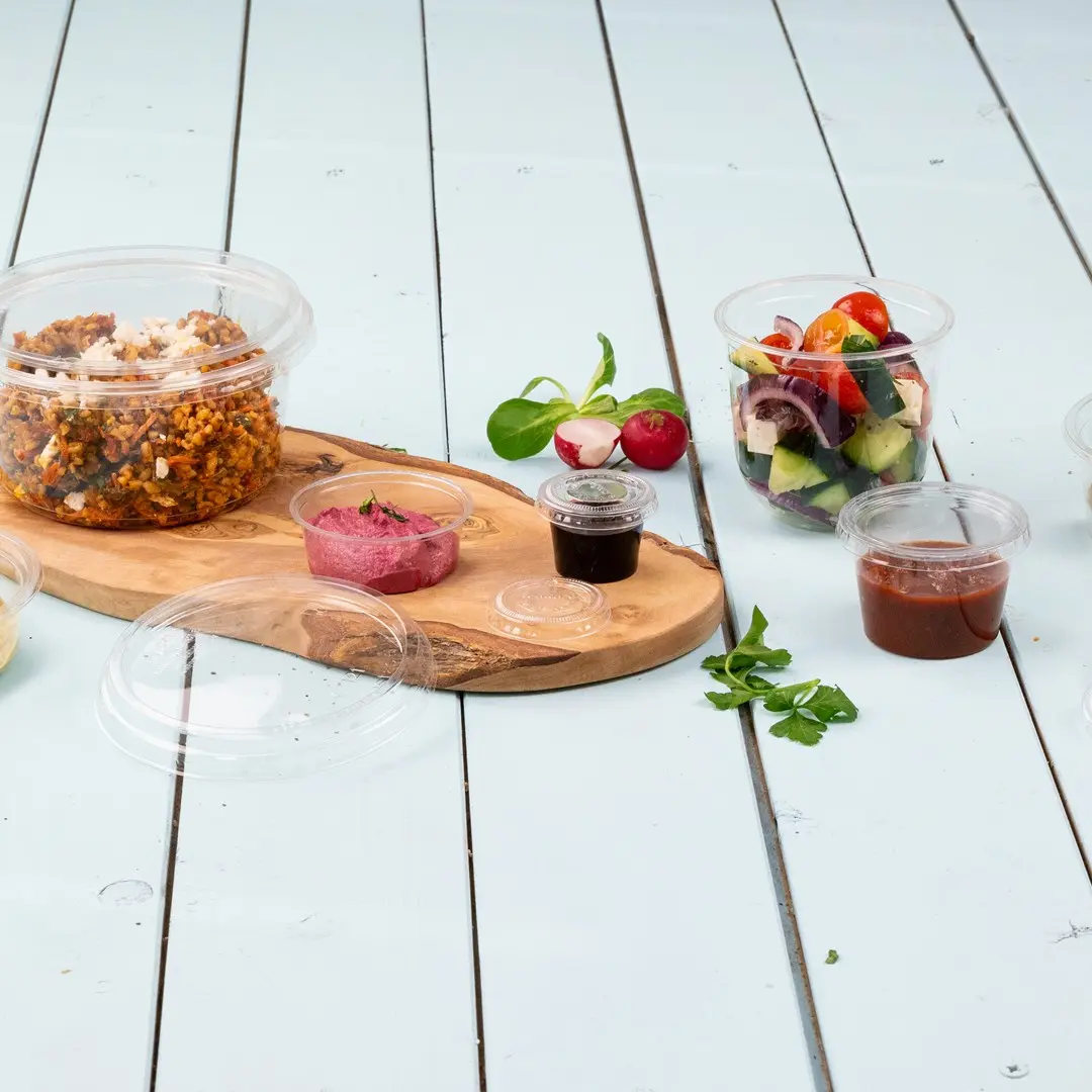 Clear plastic containers with various food items, including a salad, dips, and a grain dish, arranged on a wooden board.