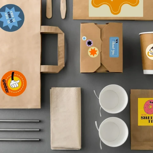 A collection of disposable food packaging including a paper bag, boxes, cups, and utensils with colorful labels and stickers.