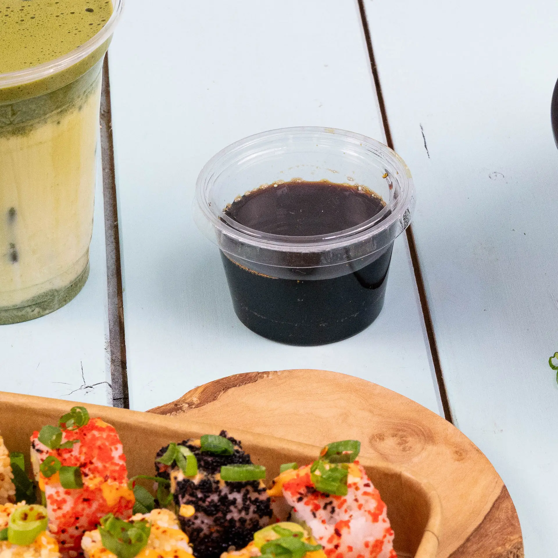 Clear round lid for portion cup with dark sauce, displayed beside beverages and sushi on a light blue surface.