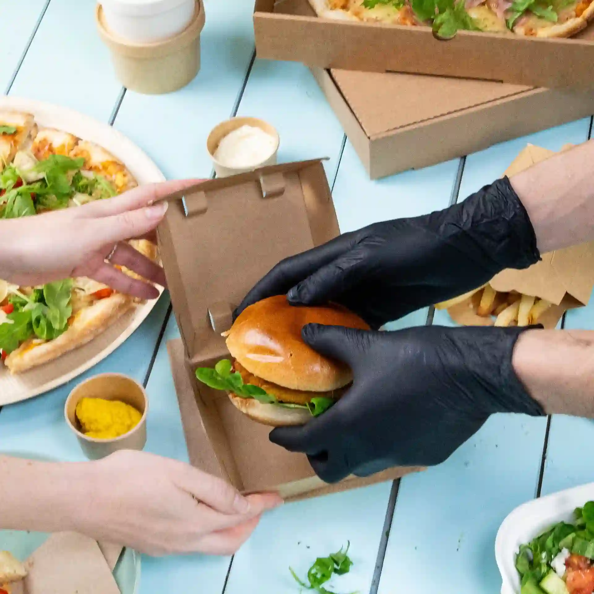A person wearing black gloves is handing a burger with lettuce to another person; pizza and fries are nearby.