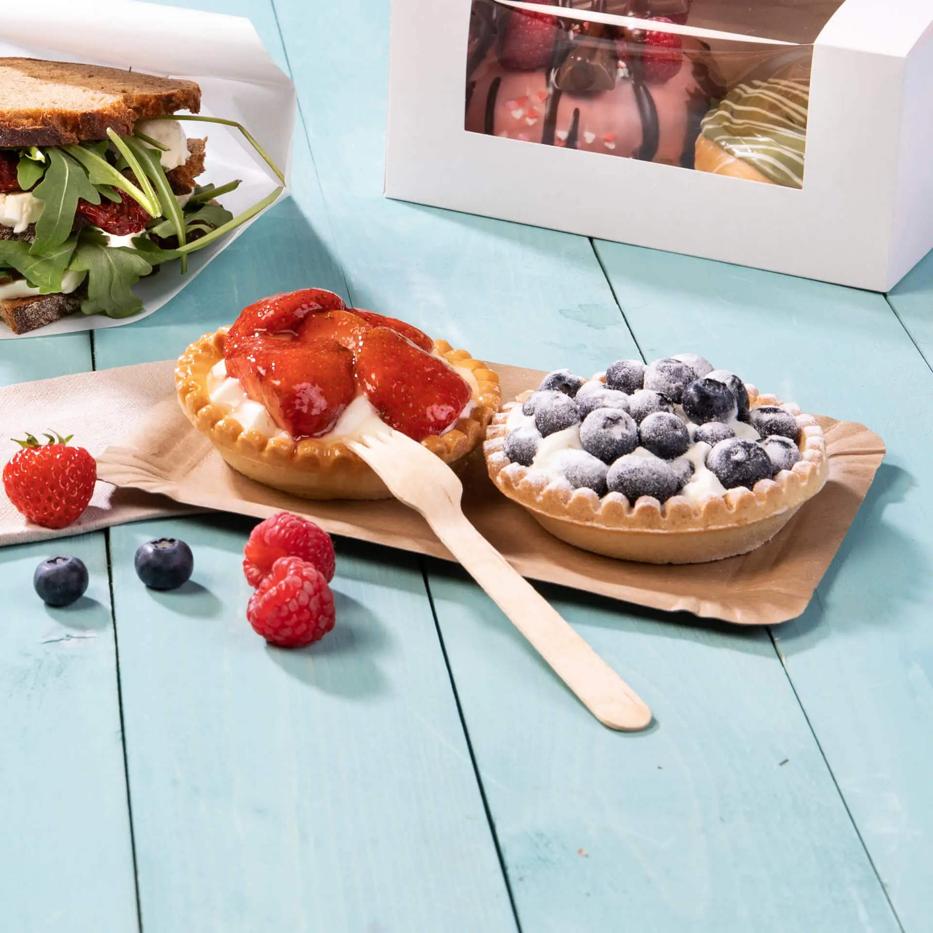 Two dessert tarts with strawberries and blueberries on a brown cardboard plate, alongside fresh berries and a sandwich.