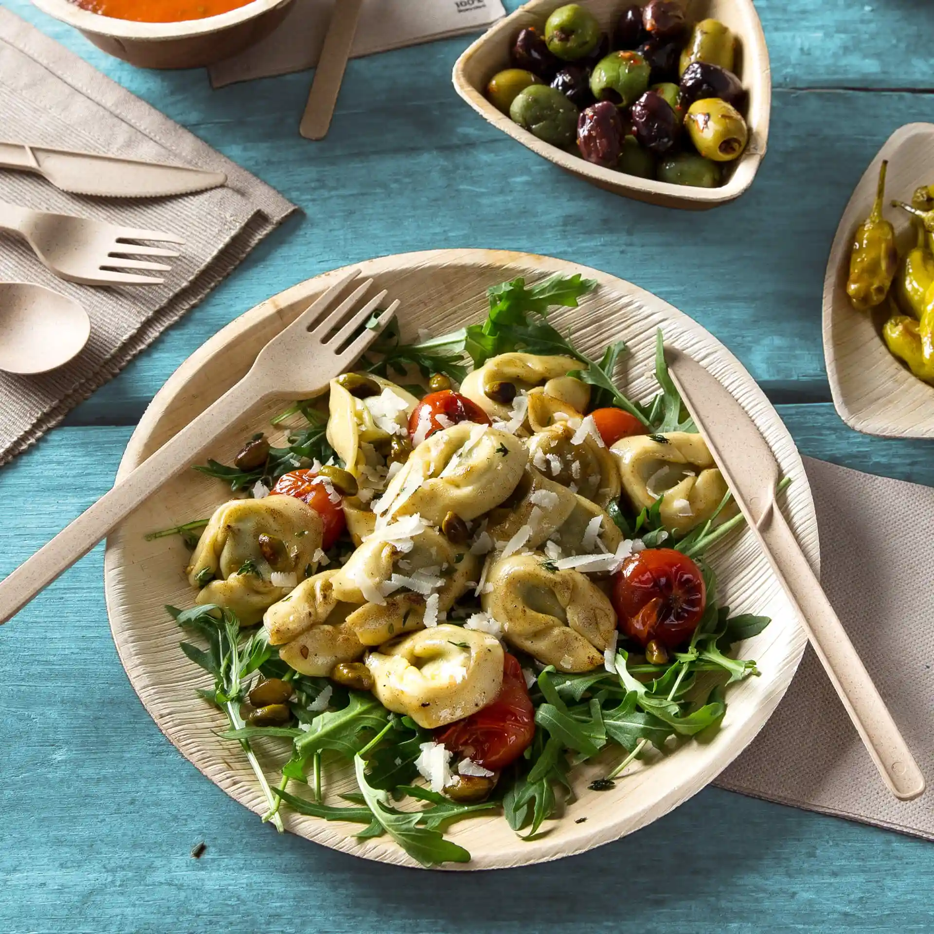Round palm leaf plate with salad, tortellini, and cherry tomatoes, accompanied by wooden utensils on a blue surface.