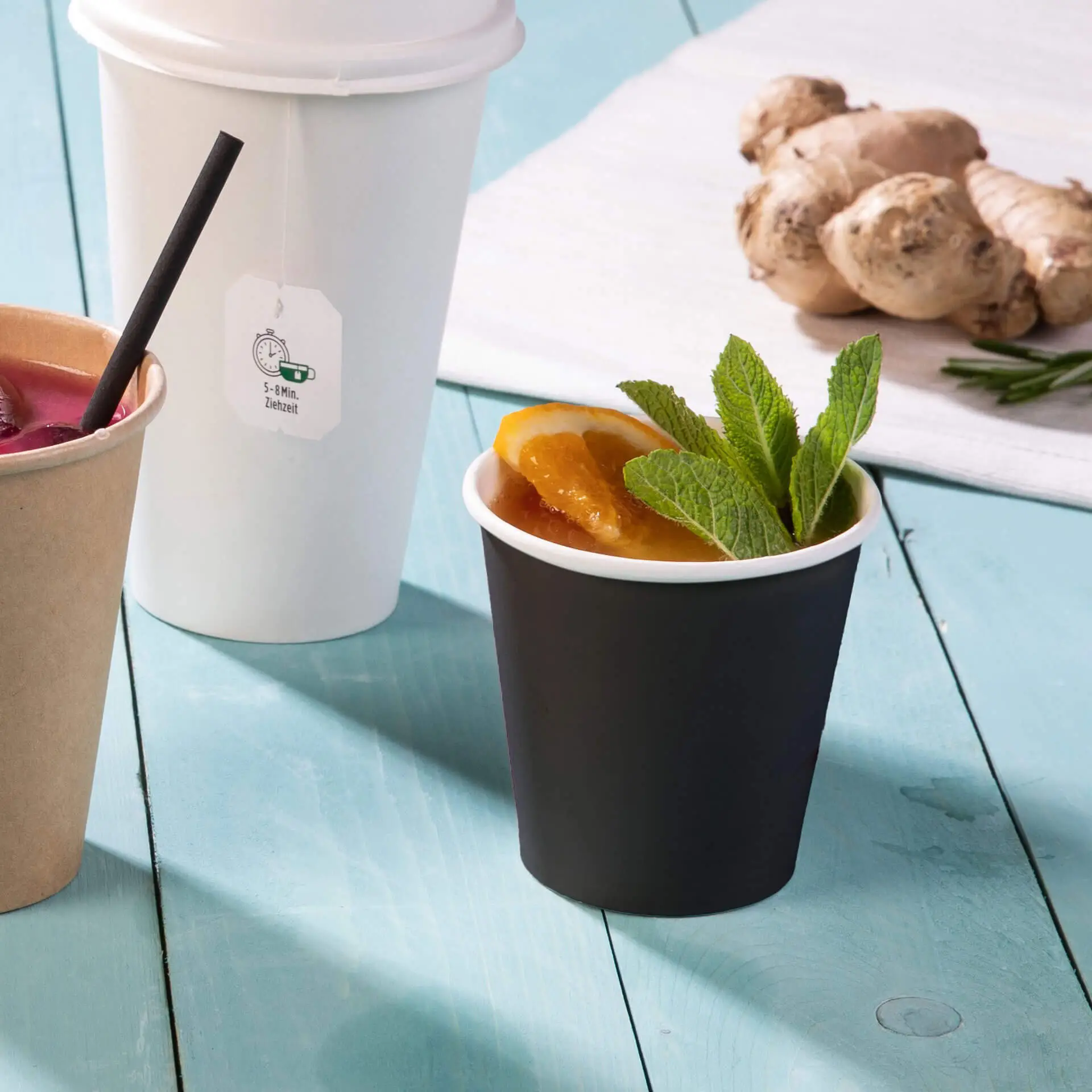 Black single-wall paper cup with PLA coating, filled with orange slices and mint leaves, alongside other cups and ginger.