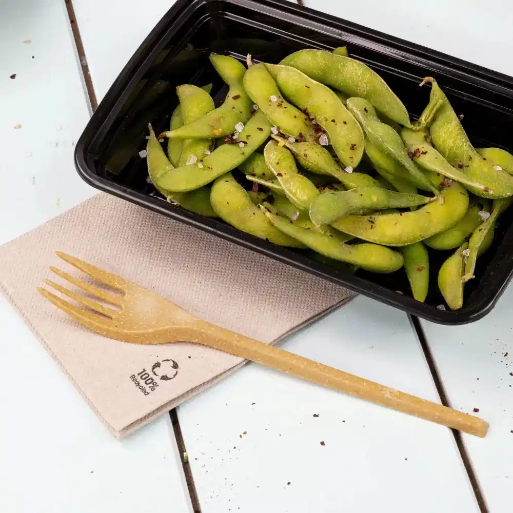 Container of edamame beans sprinkled with seasoning, beside a wooden fork on a textured napkin.