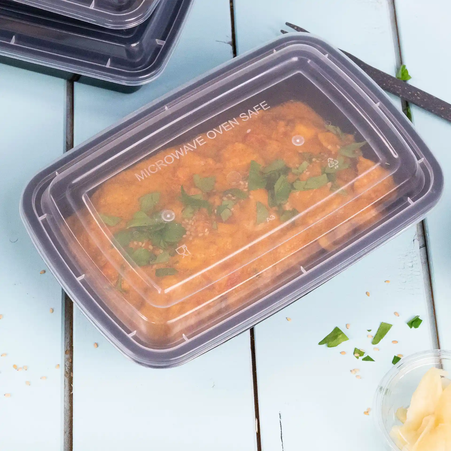 Rectangular transparent container with a reusable lid, filled with food and garnished with green herbs, on a blue surface.