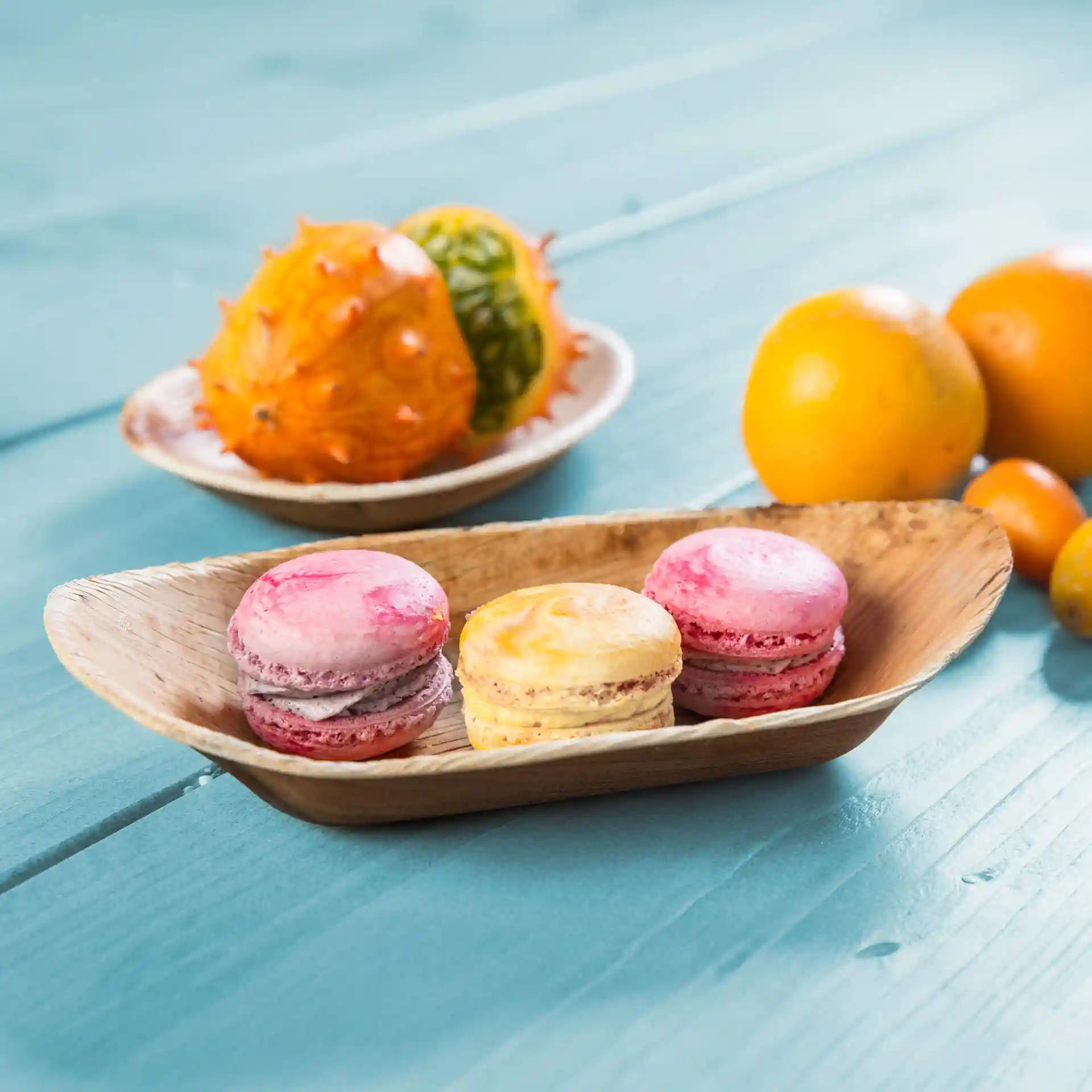 Bamboo boat-shaped dish with pink and yellow macarons, surrounded by various fruits on a blue wooden surface.