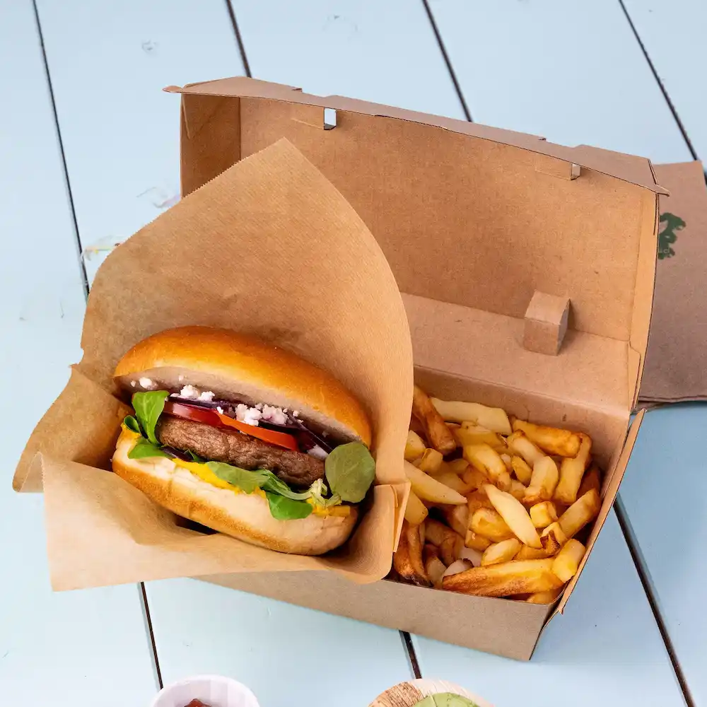 Burger in a paper wrap with fresh vegetables next to a box of French fries on a blue surface.