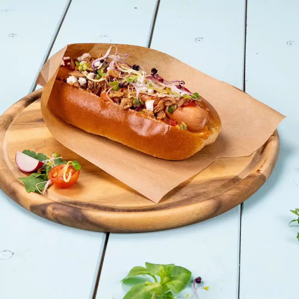 Hot dog in a soft bun topped with crispy onions, sprouts, and garnished with cherry tomatoes and radish on a wooden plate.