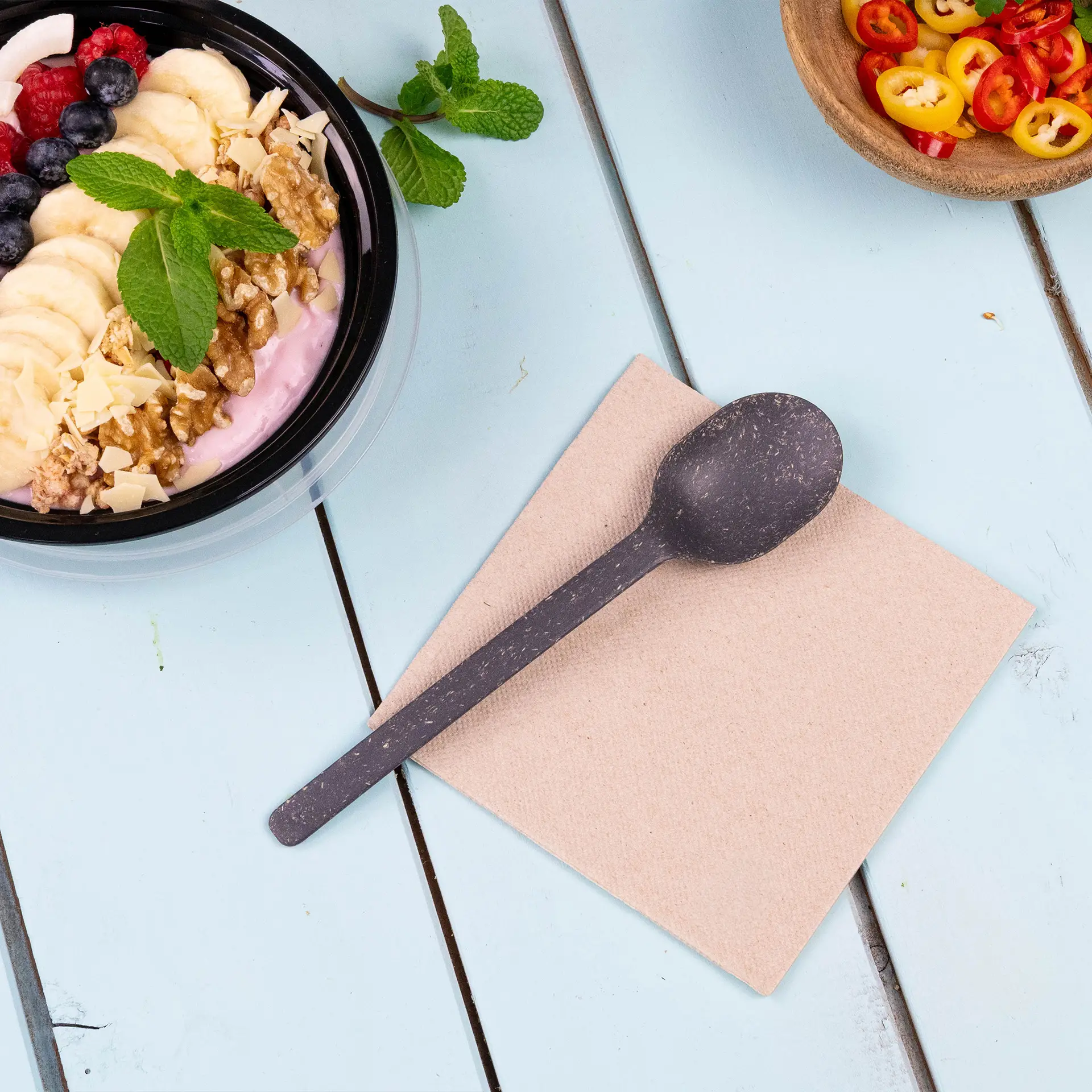 Image of a black spoon on a square unbleached napkin, with a bowl of yogurt topped with fruits and nuts in the background.