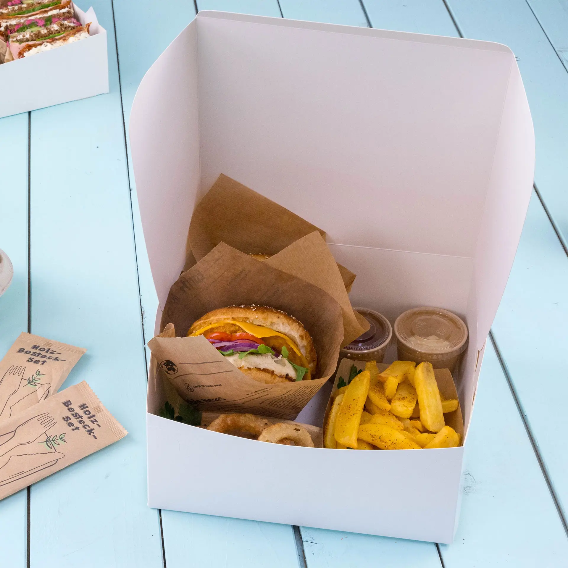 White cardboard cake box containing a sandwich, fries, and two sauce containers, with a folded lid.