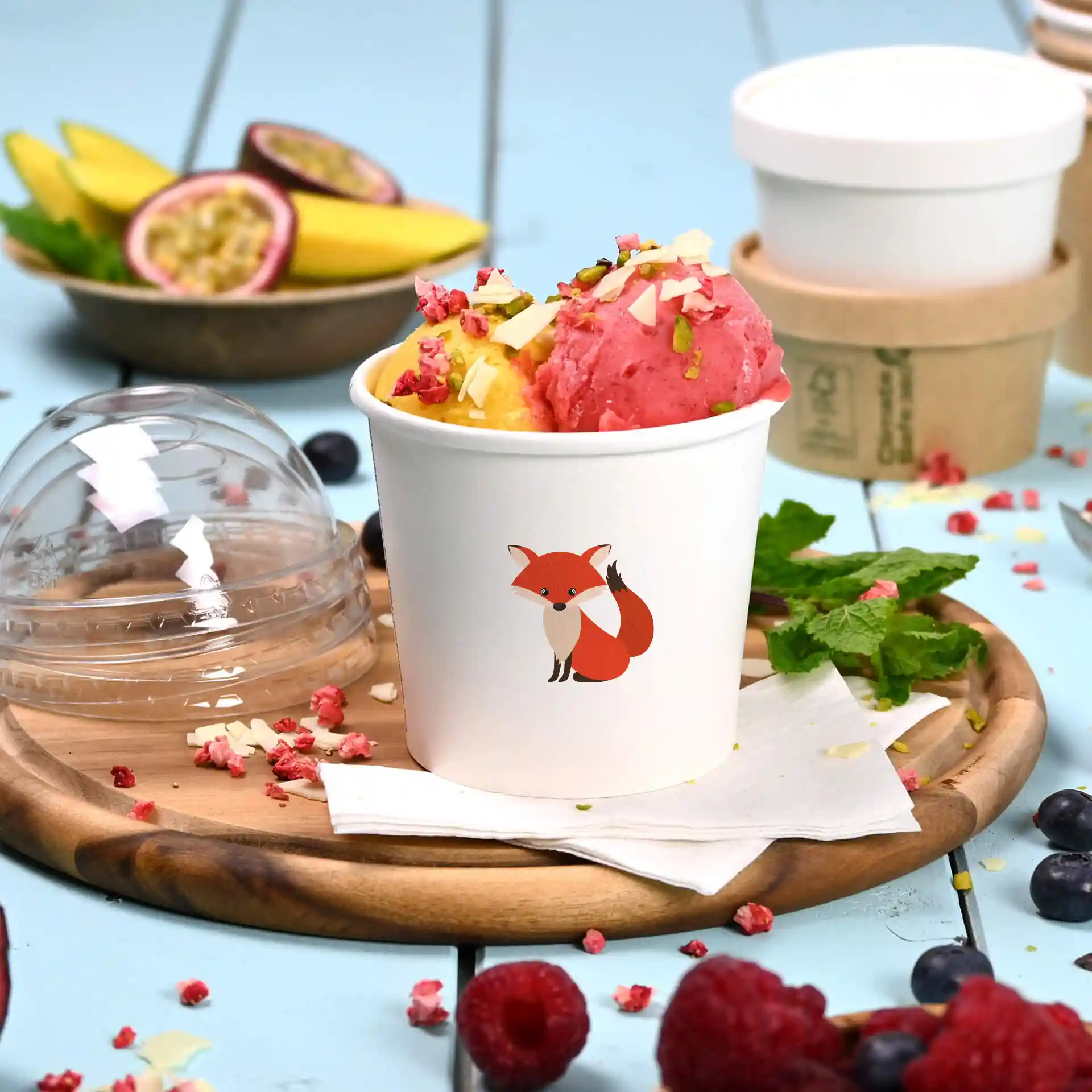 White cardboard ice cream cup with a fox illustration, filled with pink and yellow ice cream, on a wooden plate.