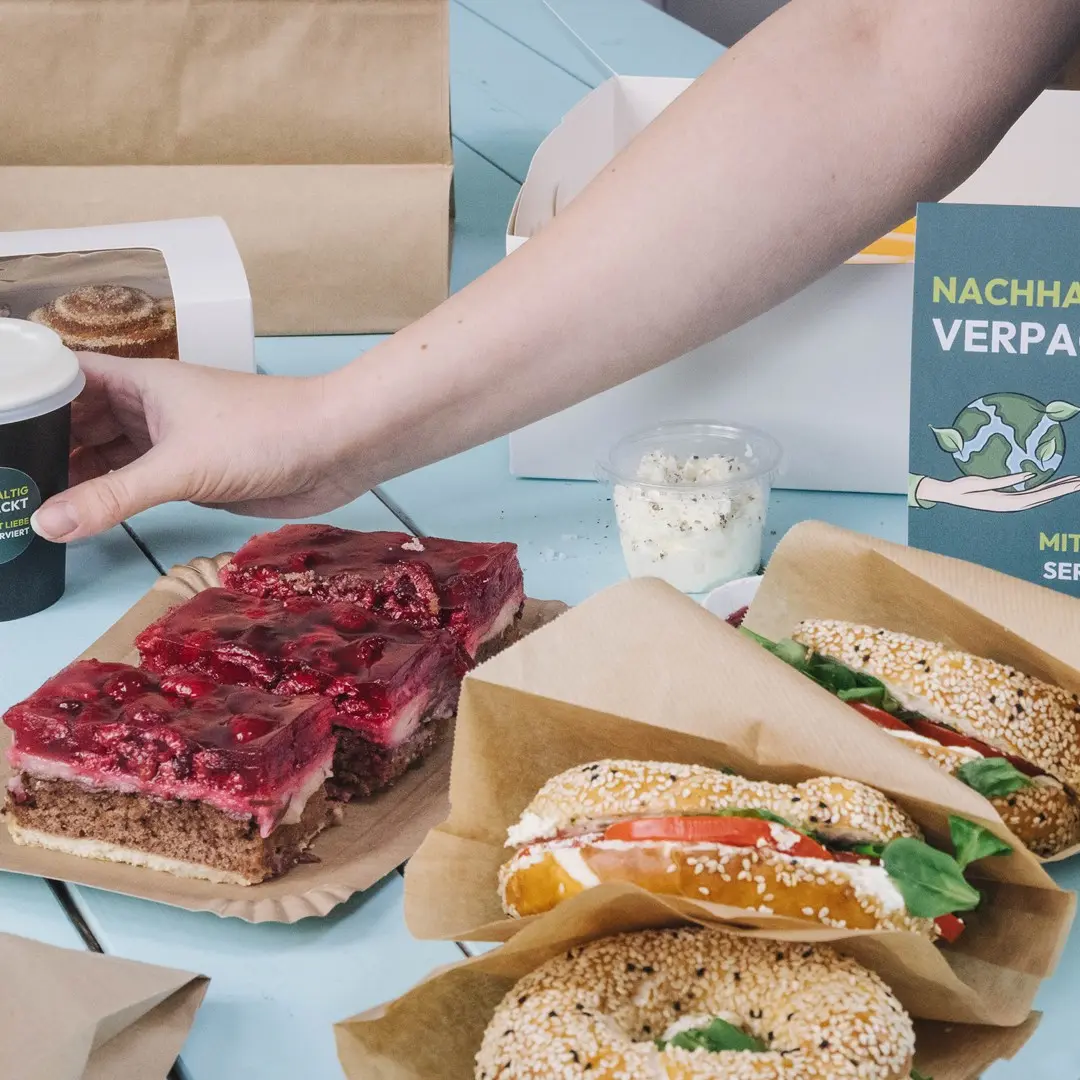 A hand reaching for a coffee cup next to a plate of layered dessert, two sesame bagels with fillings, and a container of cream.