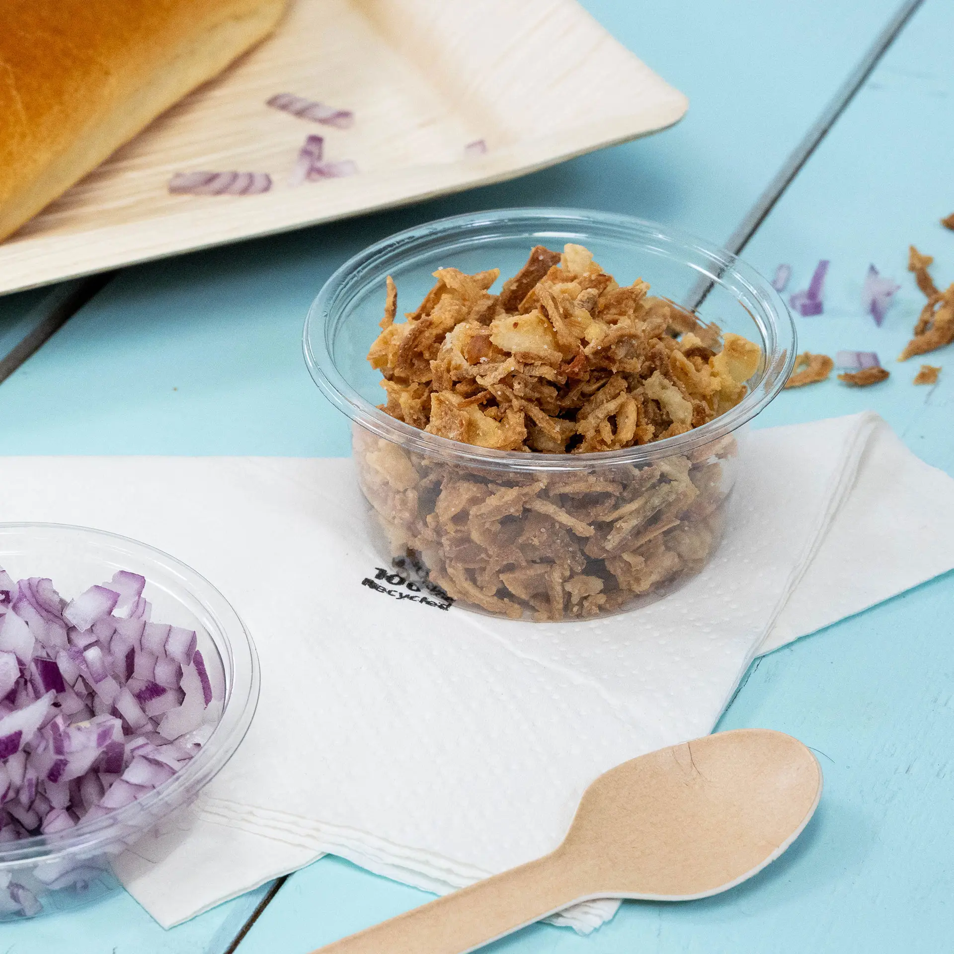 A clear cup containing crispy fried onions is placed beside chopped red onions on a napkin, with a wooden spoon nearby.