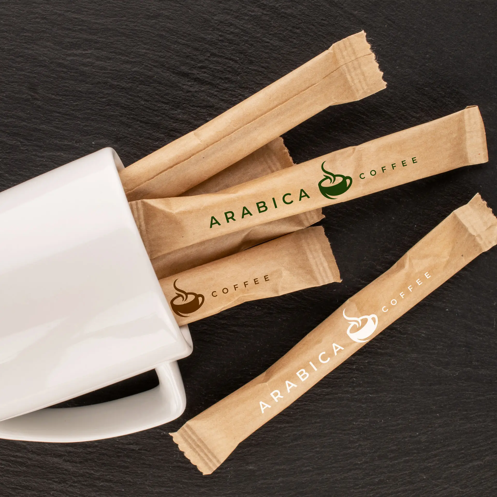 Brown organic cane sugar sticks in kraft wrappers with "ARABICA COFFEE" logo, placed next to a white mug on a dark surface.