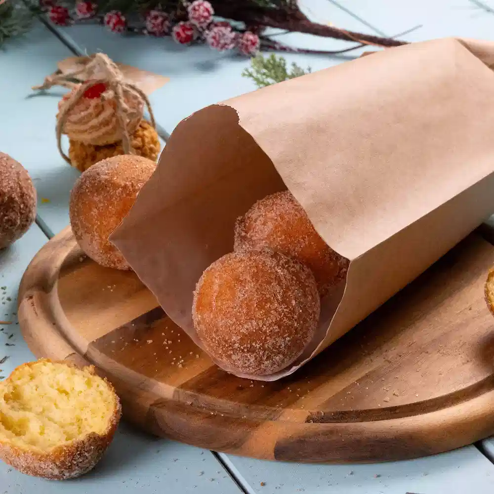 A brown paper bag contains multiple round powdered sugar donuts on a wooden cutting board, with one donut partially cut.