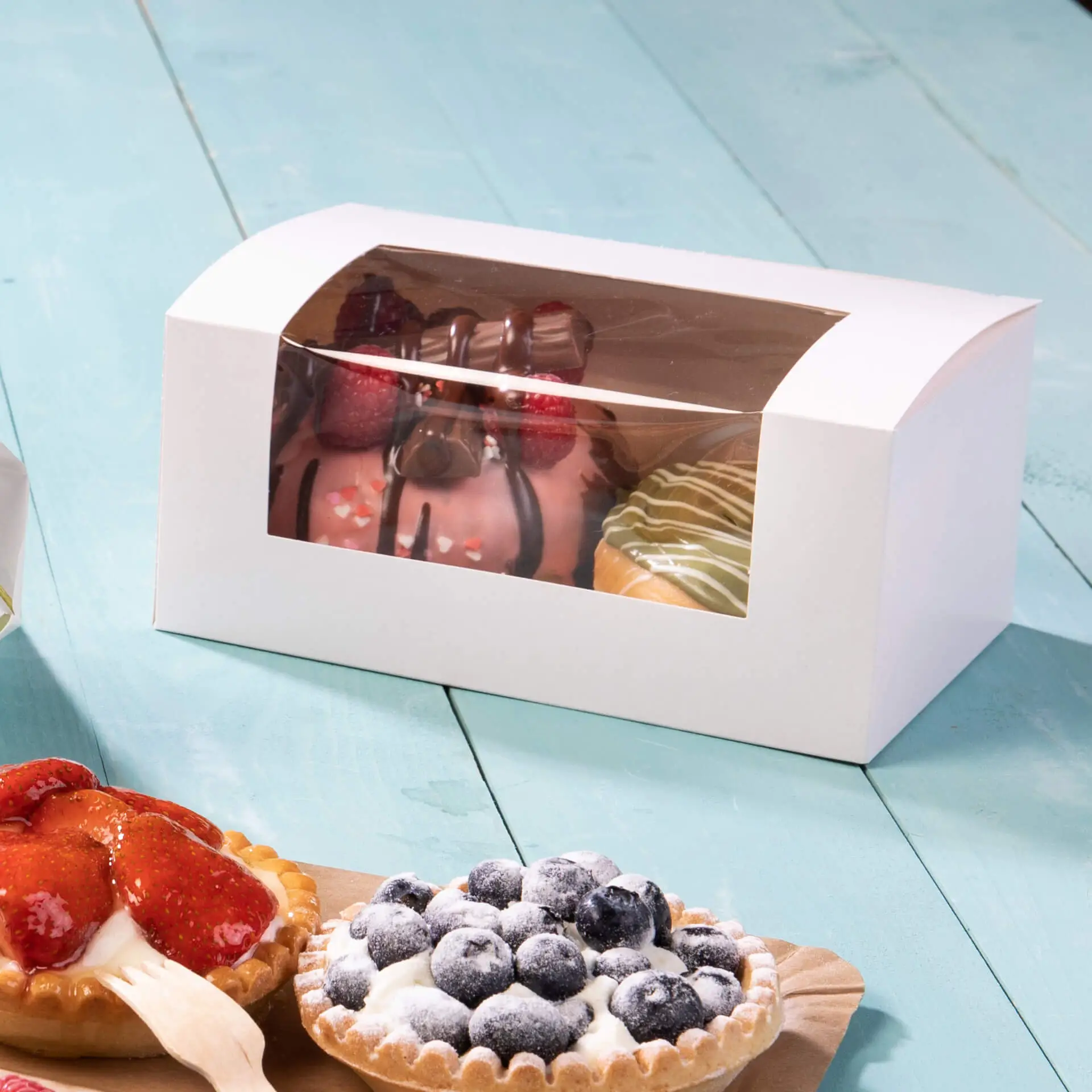 White cupcake box with a cellulose window displaying assorted pastries, including cakes and tarts, on a turquoise surface.