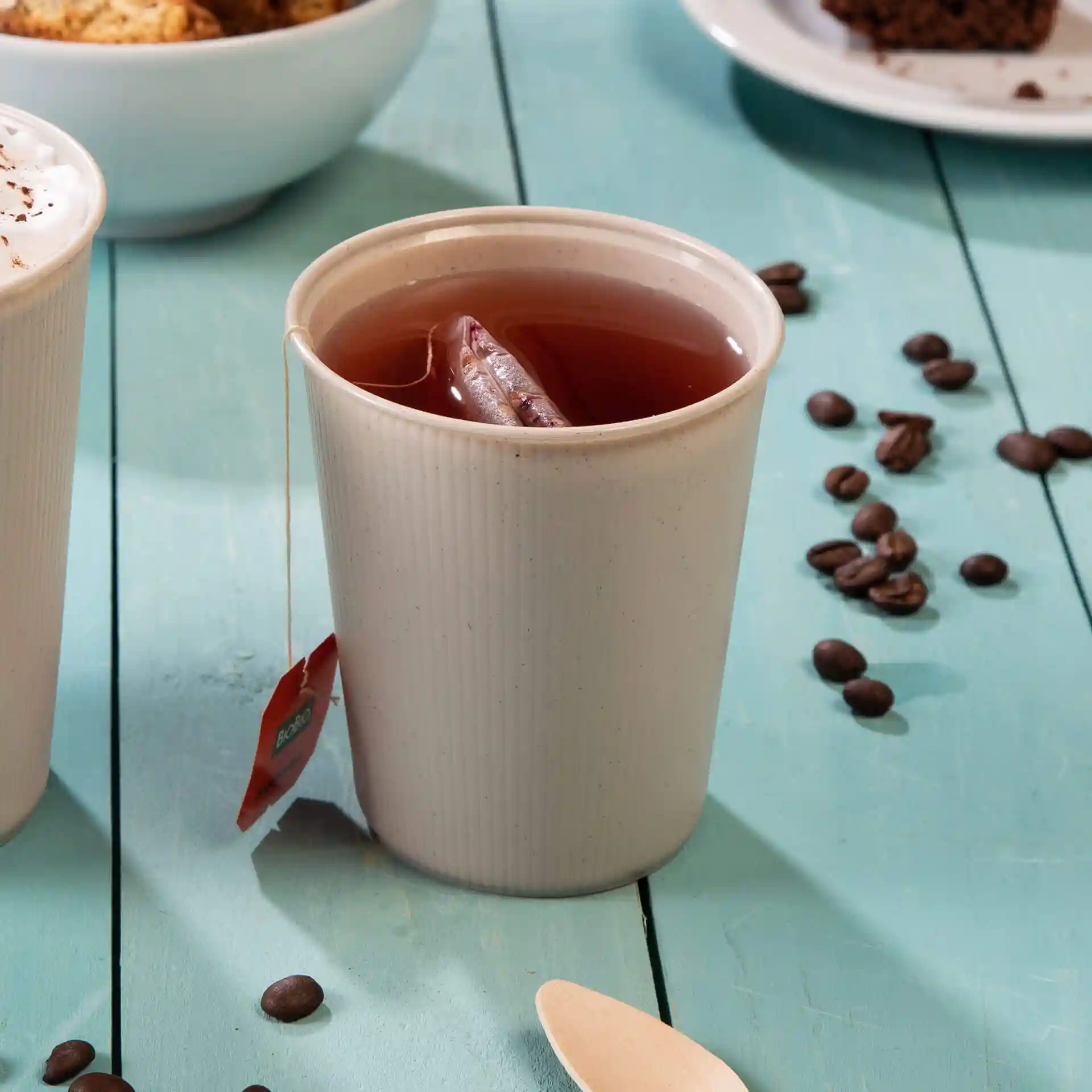 Cream white reusable coffee cup with tea bag inside, placed on a blue wooden surface with coffee beans nearby.