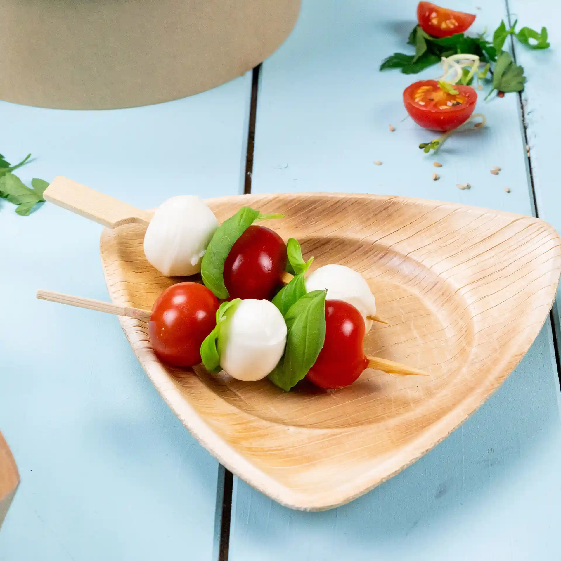 Skewers with cherry tomatoes, mozzarella balls, and basil on a wooden plate, with a light blue background.