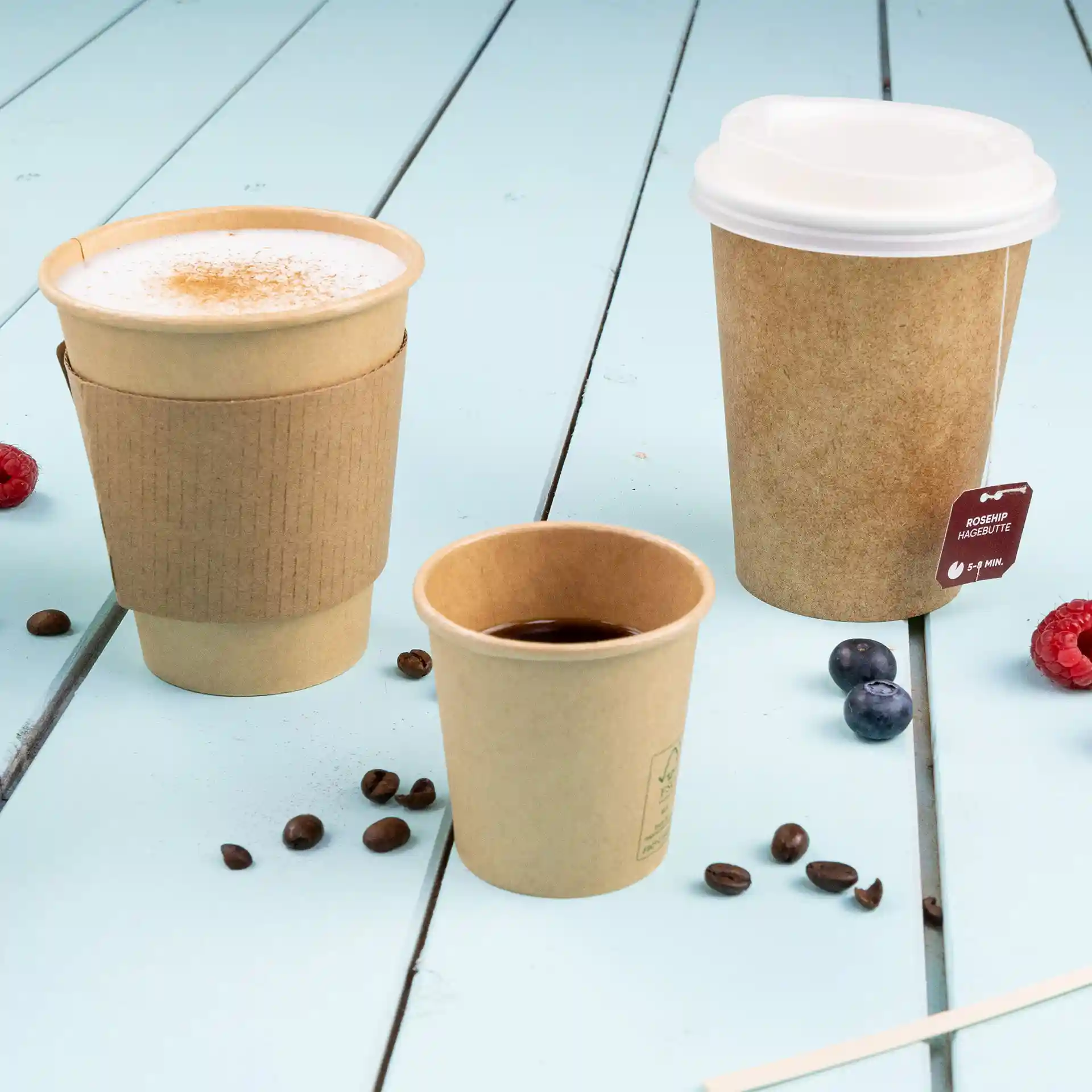 Image of three brown paper cups of varying sizes on a blue wooden surface, with coffee beans and berries scattered around.