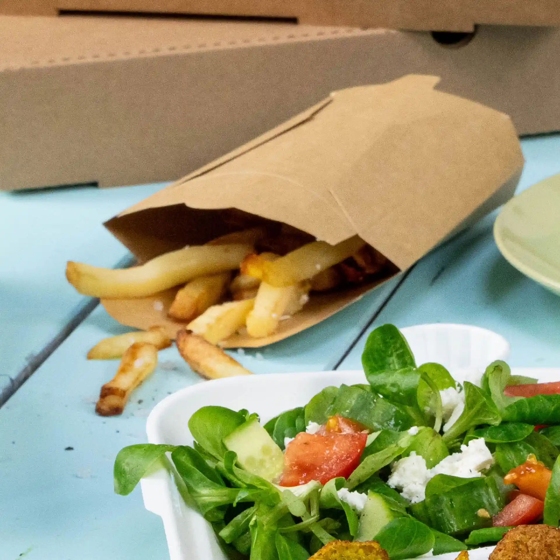 Brown cardboard chip scoop with fries spilling out, next to a salad in a white container on a blue surface.