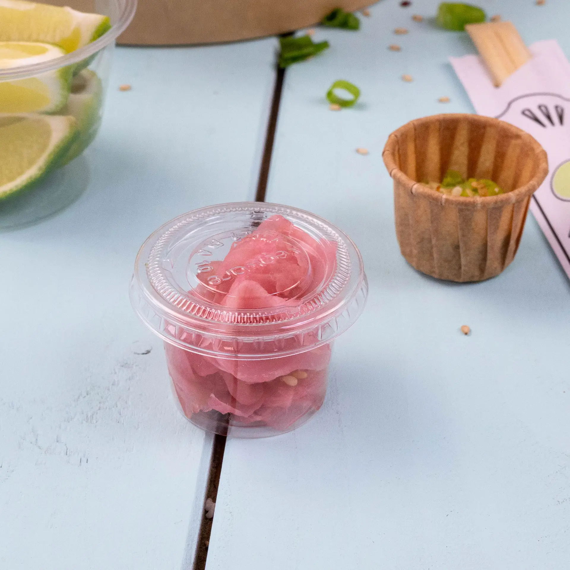 A clear plastic cup containing pink pickled ginger on a light blue surface, with a brown paper cup and lime slices in the background.