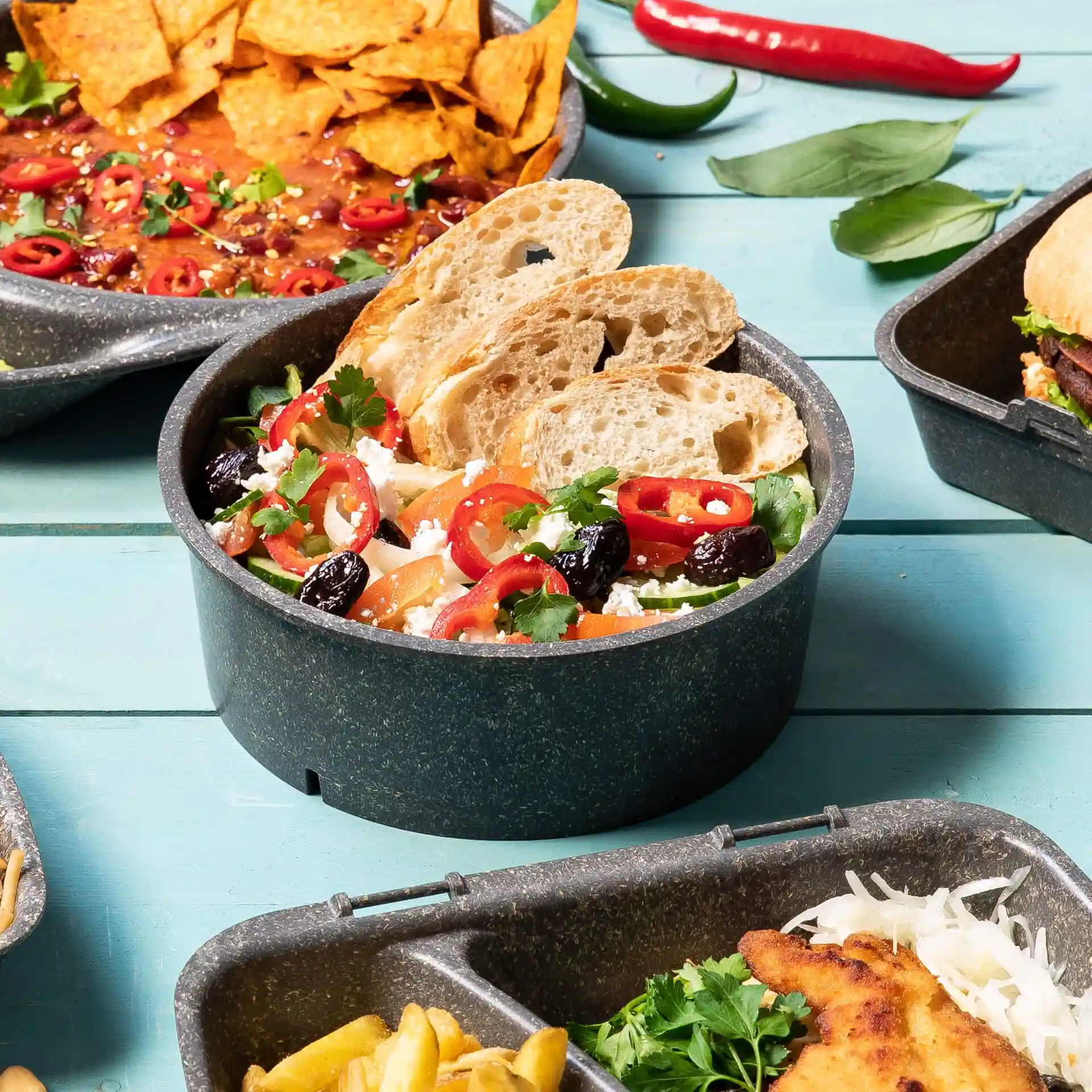 Round grey reusable bowl filled with salad, topped with sliced bread, peppers, and herbs, on a blue wooden surface.