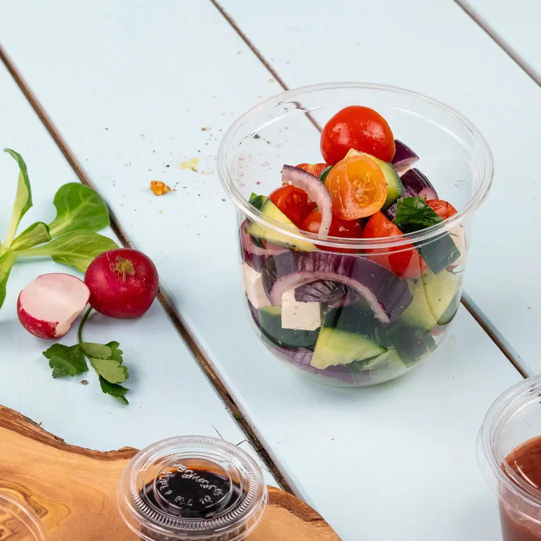 Clear plastic cup filled with mixed salad including cherry tomatoes, cucumber, red onion, and feta cheese, with radishes nearby.