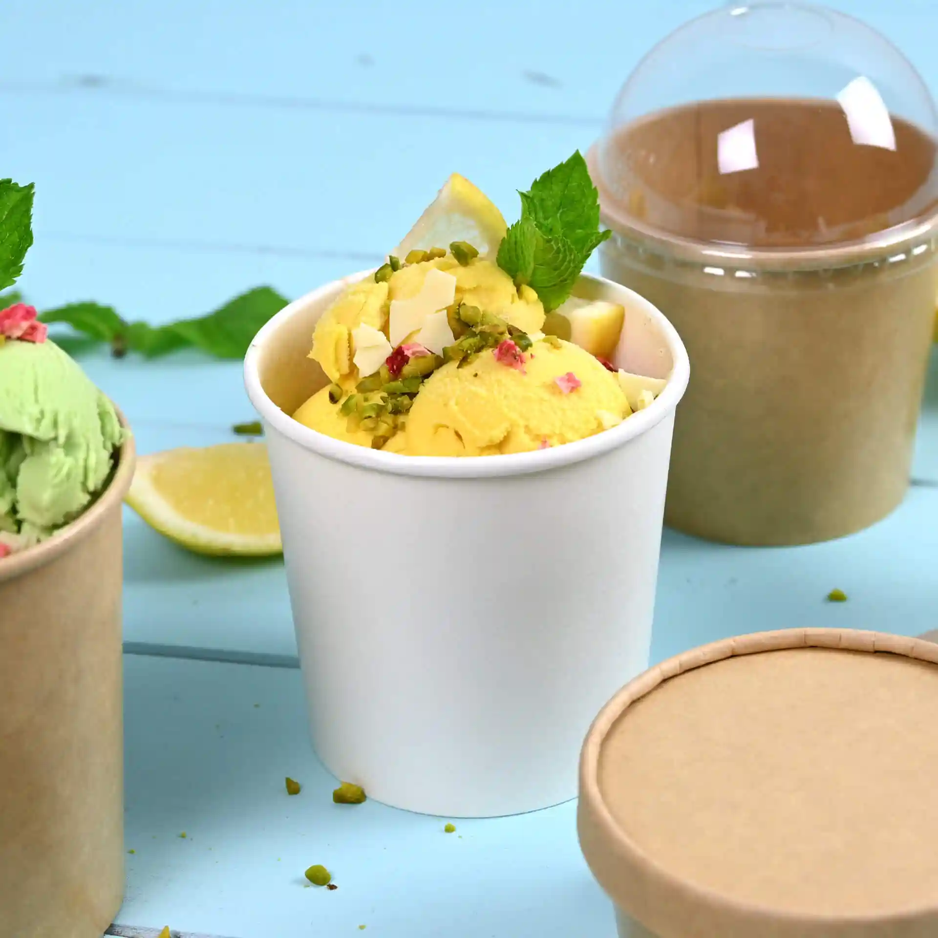 White paper ice cream tub filled with yellow ice cream, topped with mint leaves and lemon slices, with a brown tub behind.