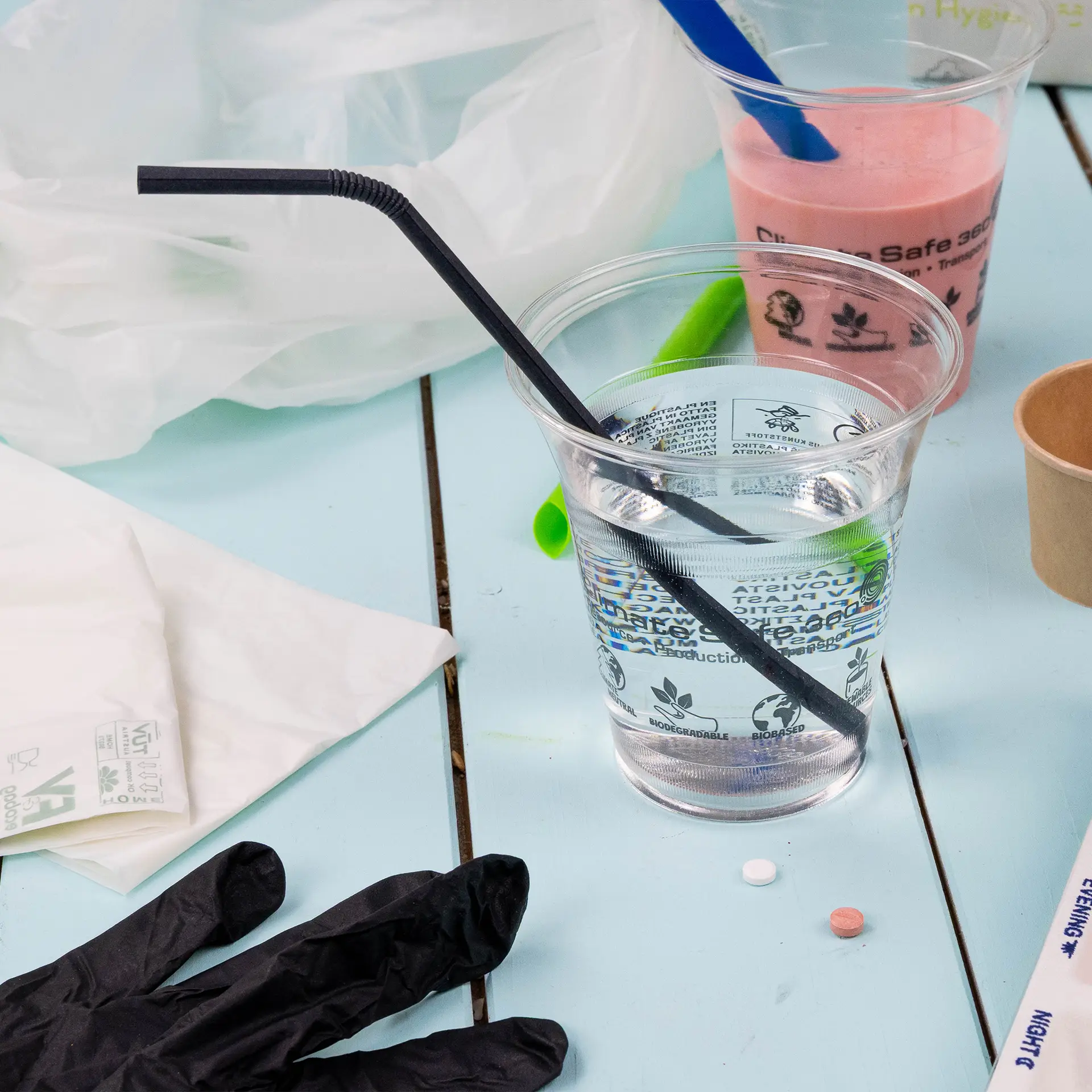 One clear cup of water with a black flexible straw, surrounded by a black glove, a pink cup, and various other items.