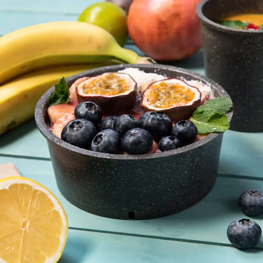 A black bowl filled with fruit, including blueberries and passion fruit, surrounded by bananas and a lemon slice.