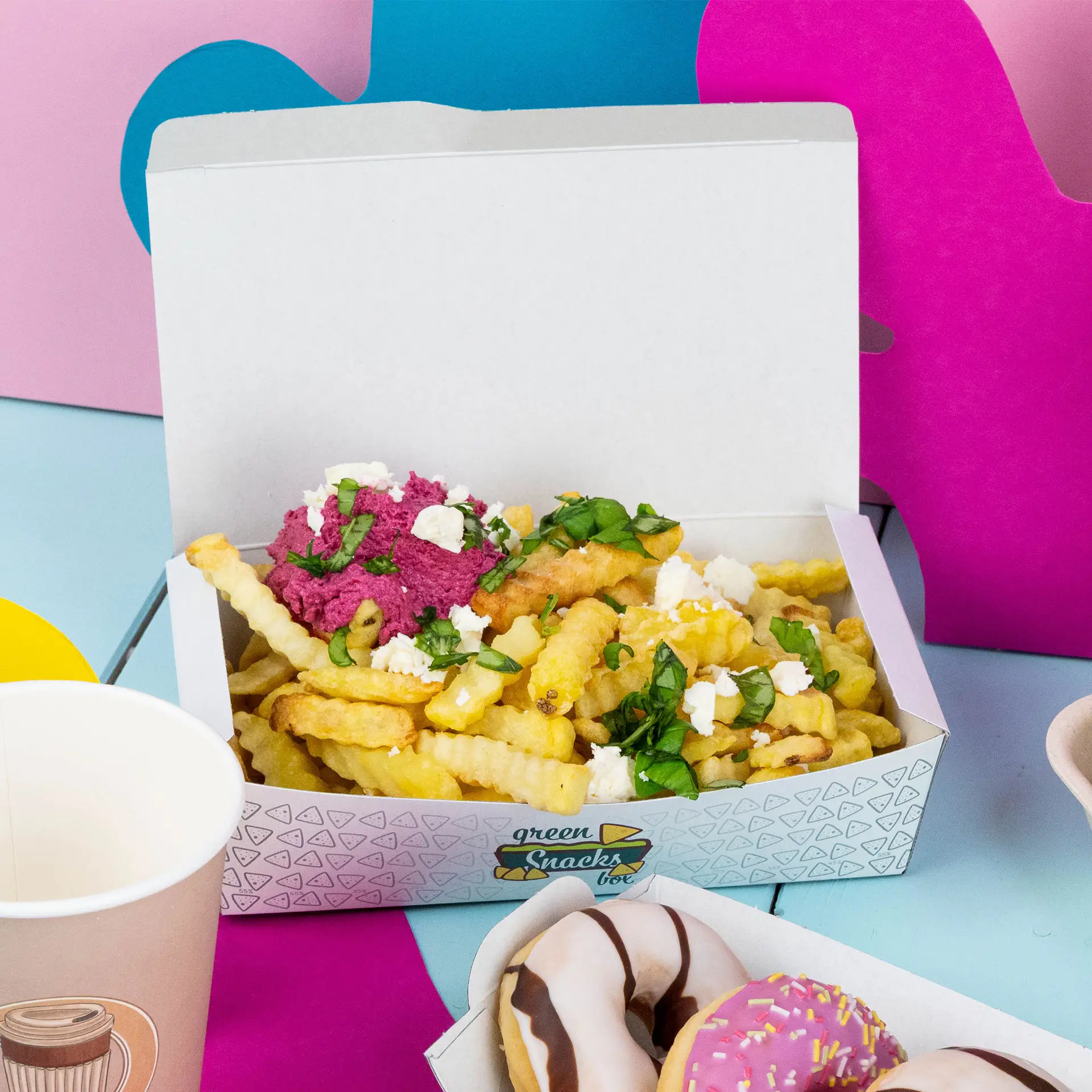 White snack box containing crinkle-cut fries topped with pink dip, herbs, and cheese, with donuts and a cup nearby.