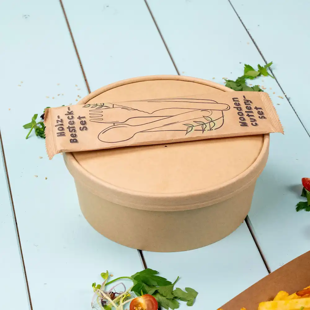 Brown container with a lid and a packaged wooden cutlery set on top, with green herbs and salad elements nearby.