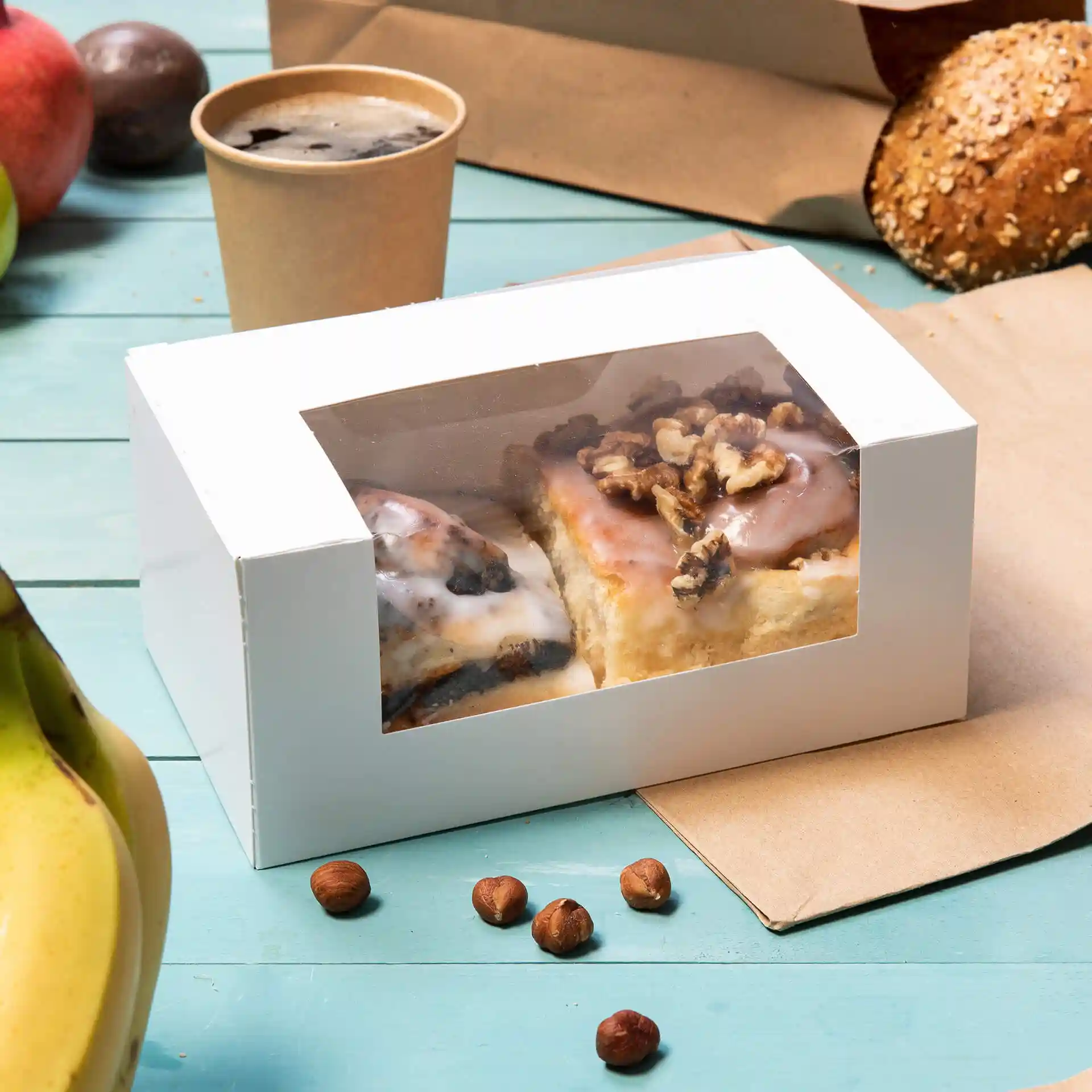 White pastry box with a cellulose window containing two pastries, surrounded by a banana, hazelnuts, and a coffee cup.