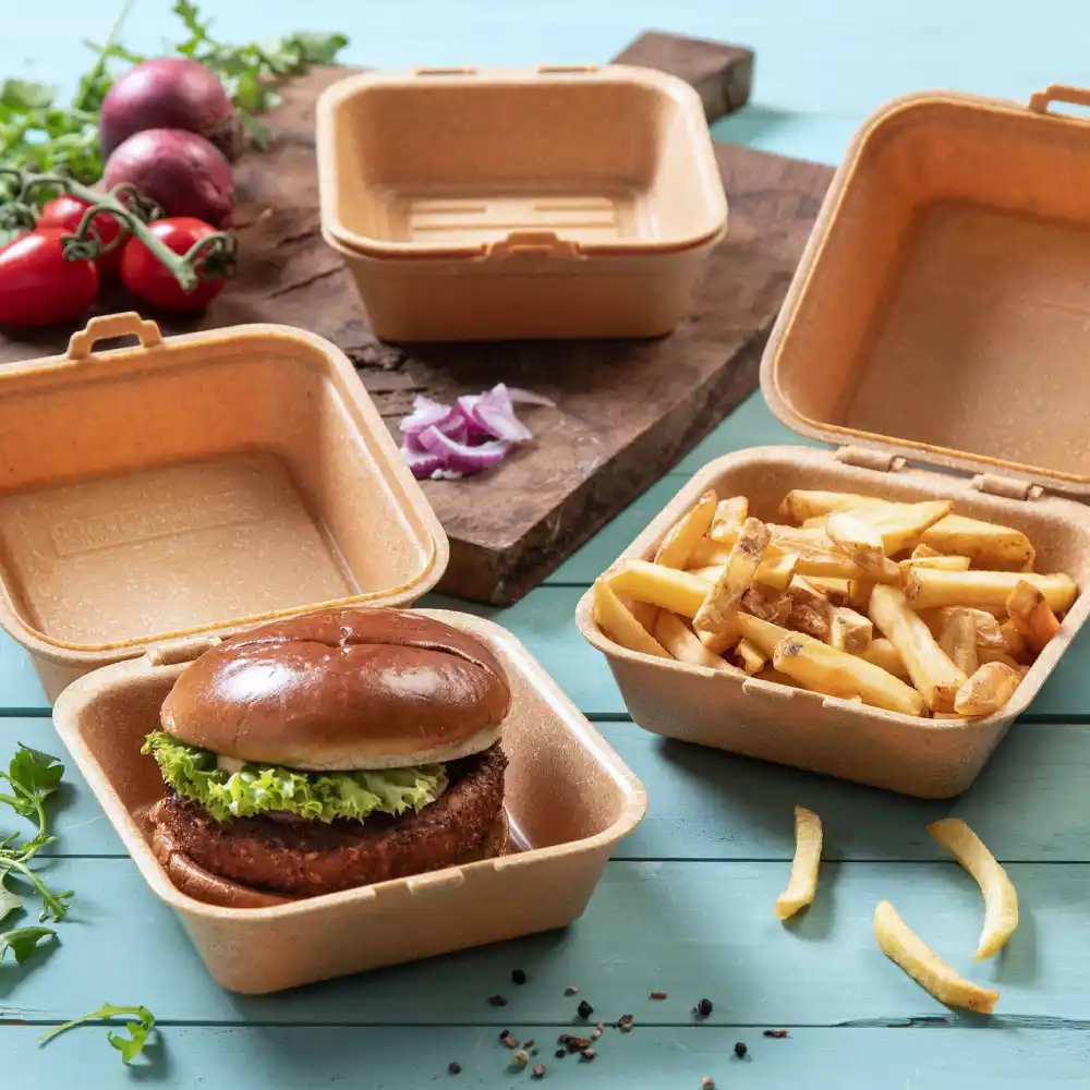 Container with a burger topped with lettuce, next to a container with fries, on a wooden surface with vegetables nearby.