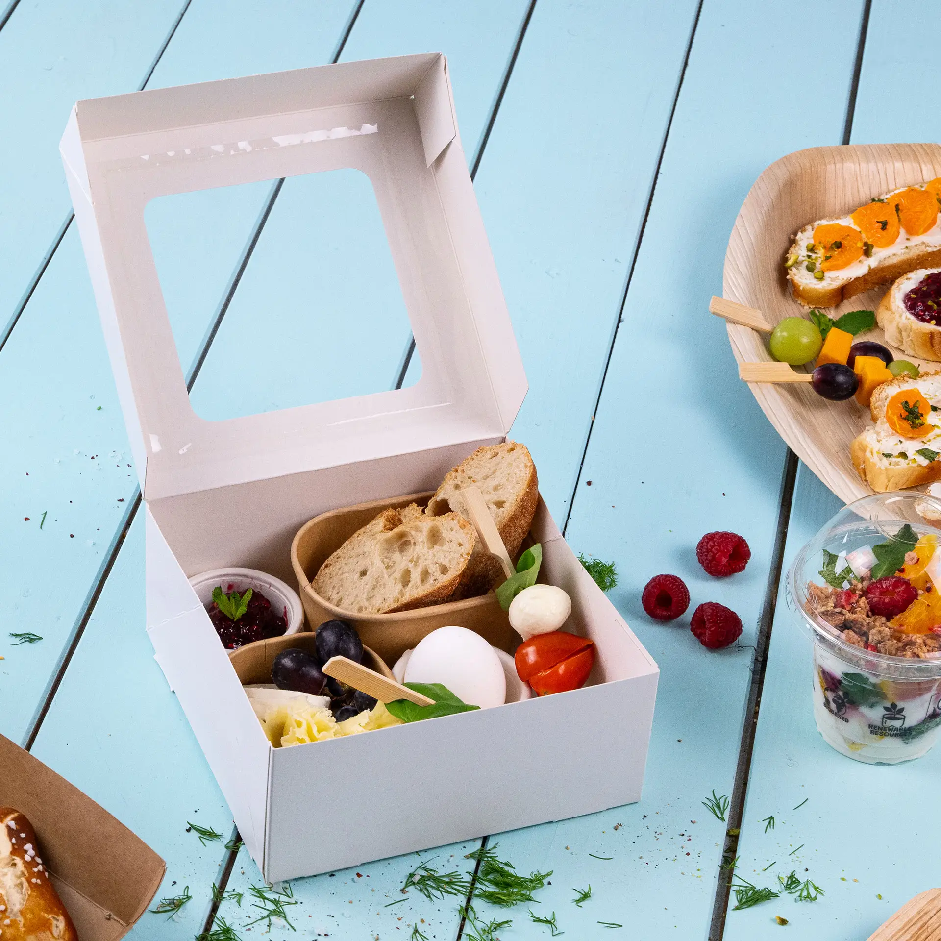 White cupcake box with a cellulose window, containing bread, cheese, grapes, an egg, and a small cup of jam.