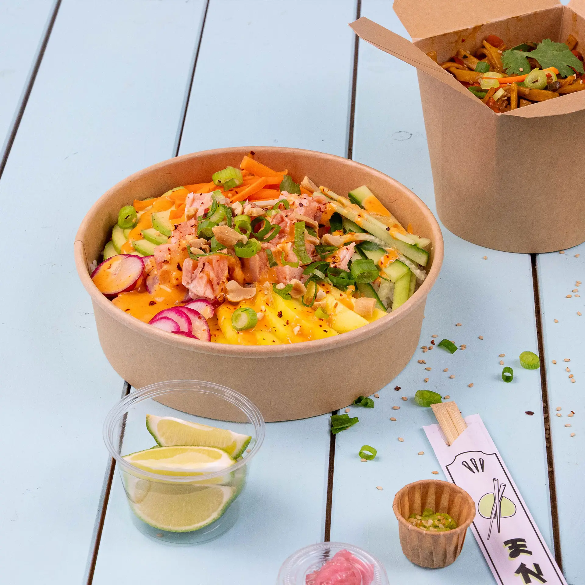 Brown paper salad bowl filled with colorful vegetables, topped with salmon and green onions, alongside lime slices.
